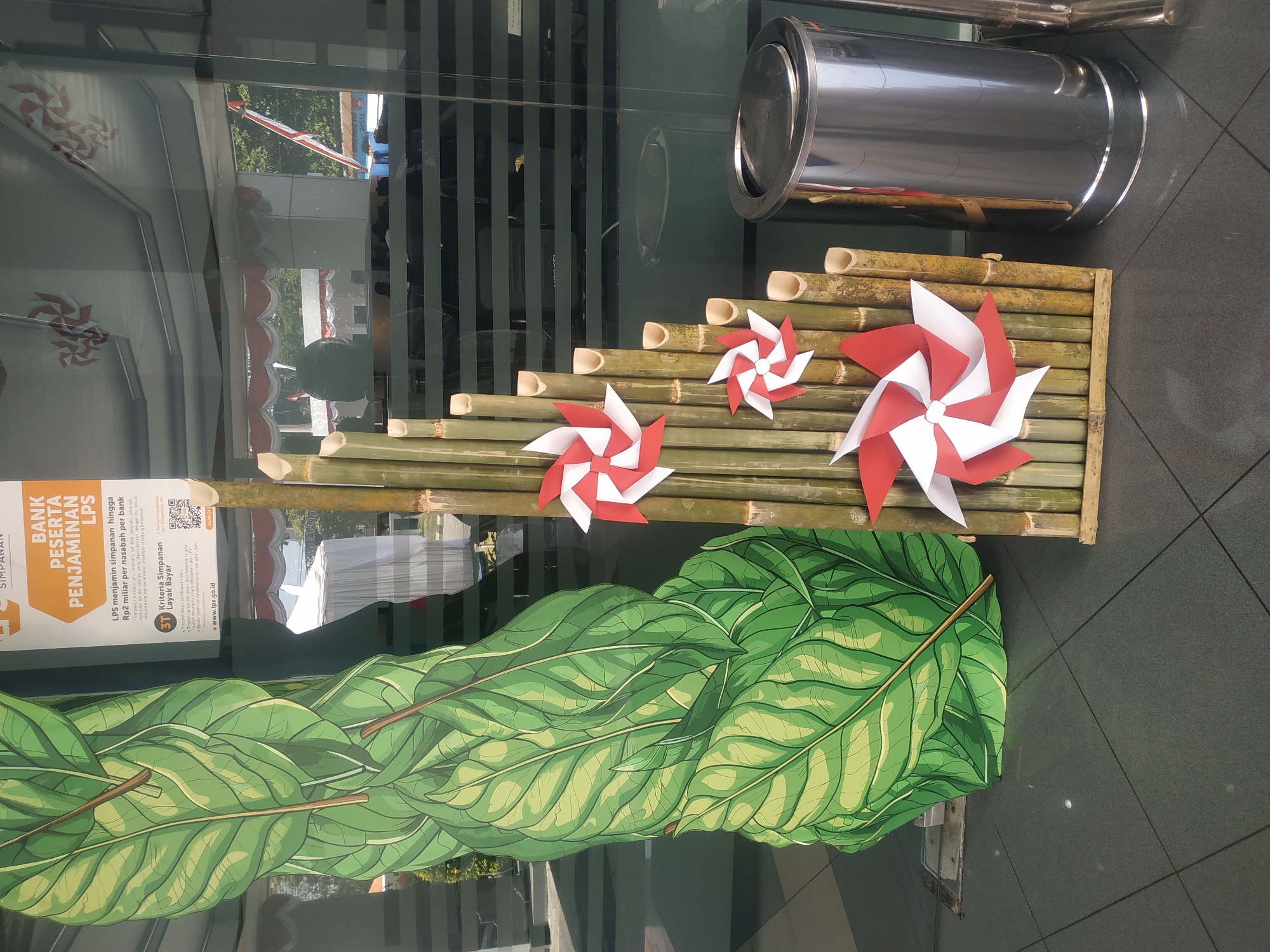 The image shows an outdoor scene in Jember, Indonesia, likely in front of a building with a glass facade. A large, stylized green leaf decoration stands to the left. To its right, a vertical arrangement of bamboo poles is decorated with red and white pinwheels. The bamboo structure has a stepped appearance. In the far right, a shiny silver trash can is visible. The reflection in the glass facade shows a building interior with tables and chairs, a ceiling fan, and red and white draped decorations. Another reflection shows an outdoor scene with trees and a flagpole with the Indonesian flag. The ground is paved with dark grey tiles. There are no people clearly visible in the image. The overall impression is of a decorative display, possibly for a public building or event.