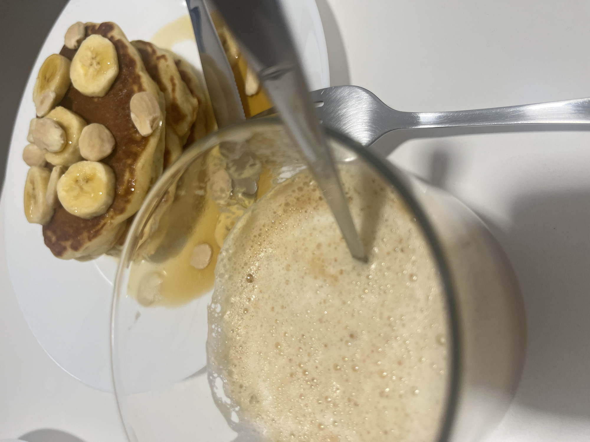 A close-up shot shows a white plate with a stack of pancakes topped with sliced bananas and nuts, drizzled with syrup. Adjacent to the plate is a glass filled with a light-brown, frothy beverage. A spoon and fork rest on the white surface beside the plate and glass. The scene appears to be indoors, likely a breakfast setting. The location is unknown, and there is no confirmed relationship to Mogán, Spain. There are no people visible in the image, and no actions or interactions are occurring beyond the arrangement of the food and drink items.