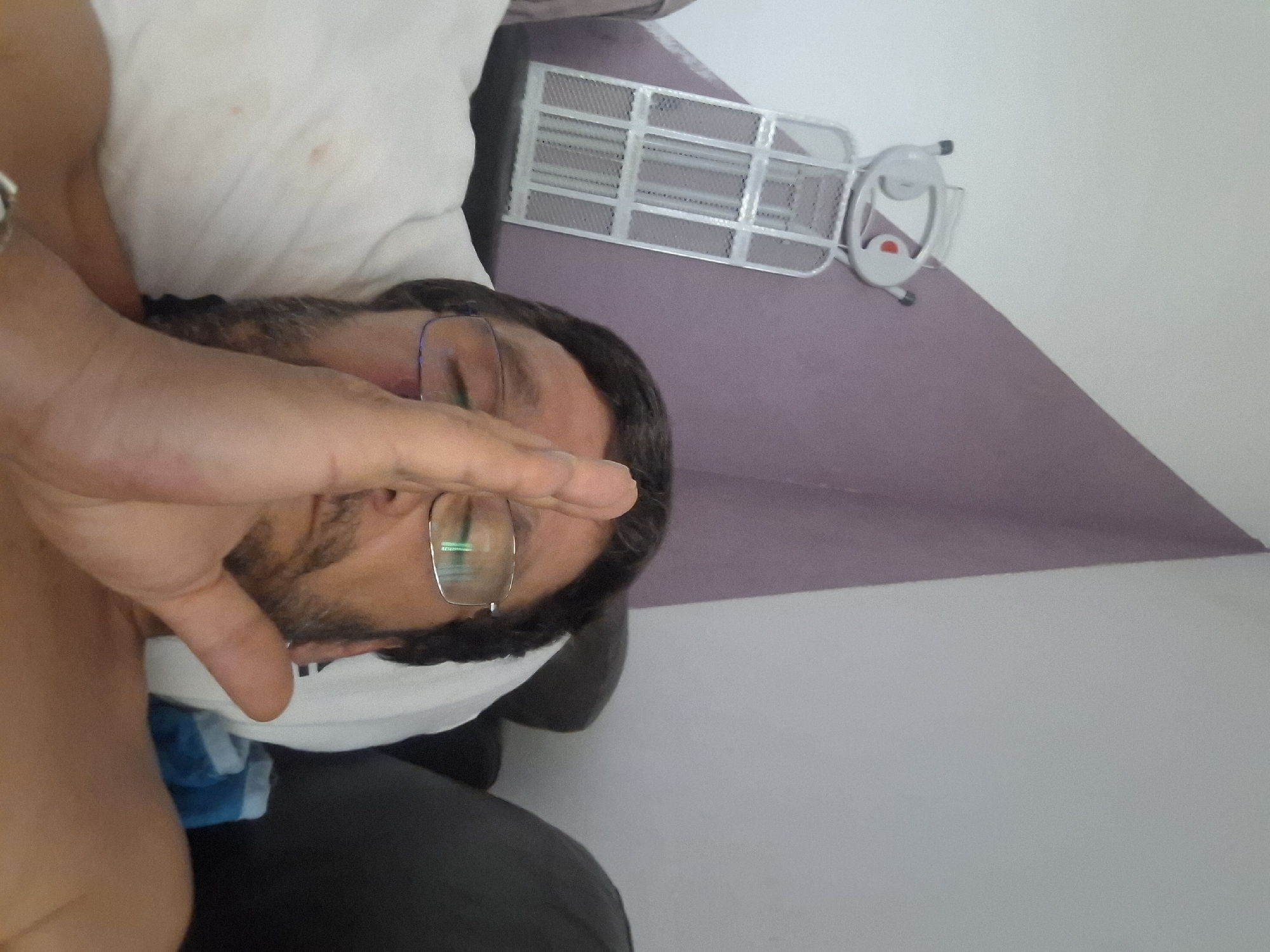 A man with glasses and a beard, lying on a pillow, is captured in a close-up selfie. His hand is raised, partially obscuring his face. In the background, a metallic object with a basket-like structure and a circular top with two ear-like protrusions is visible against a purple and white wall. The scene appears to be indoors, likely in a residential setting within Yerocham, Israel, given the provided context.