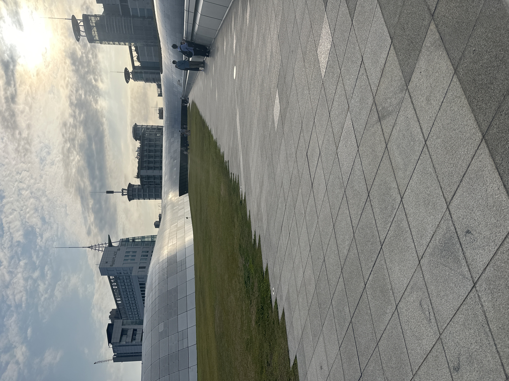 The image depicts the Dongdaemun Design Plaza (DDP) in Seoul, South Korea. The foreground is dominated by a gray, tiled walkway with a grid pattern. Adjacent to the walkway is a strip of green grass. In the background, modern buildings and skyscrapers are visible against a bright, cloudy sky. Several people are standing or walking along the walkway. The overall composition suggests an architectural and urban environment.