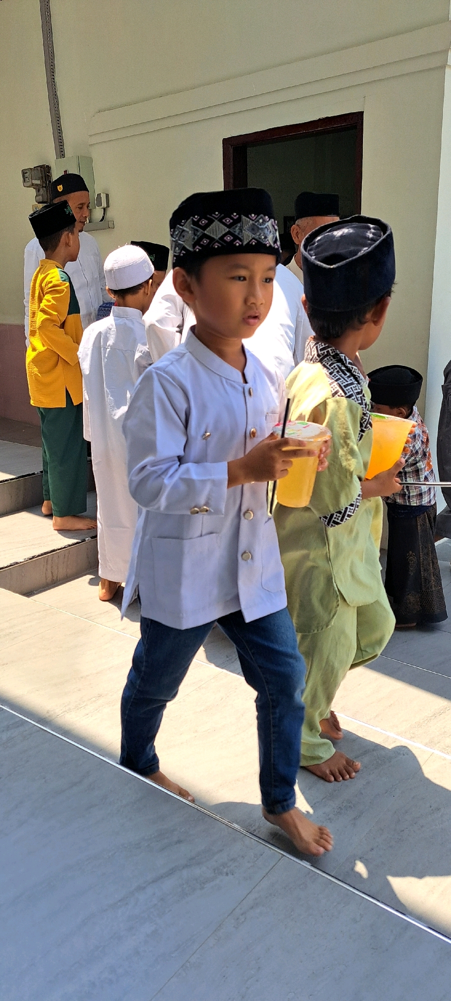 In the city of Sumber Gedang, Indonesia, a group of young boys, dressed in traditional Islamic attire, are seen walking outdoors on a bright, sunny day. The boy in the foreground, wearing a white long-sleeved shirt and blue jeans, is holding a yellow drink in a plastic cup with a straw. To his right, another boy in a light green outfit also holds a similar yellow drink. Several other boys are visible in the background, some in white robes and skullcaps, while others are partially obscured. The scene appears to be on a paved area with steps, suggesting a pathway or entrance to a building. The overall atmosphere is one of community and celebration, possibly related to a religious event.