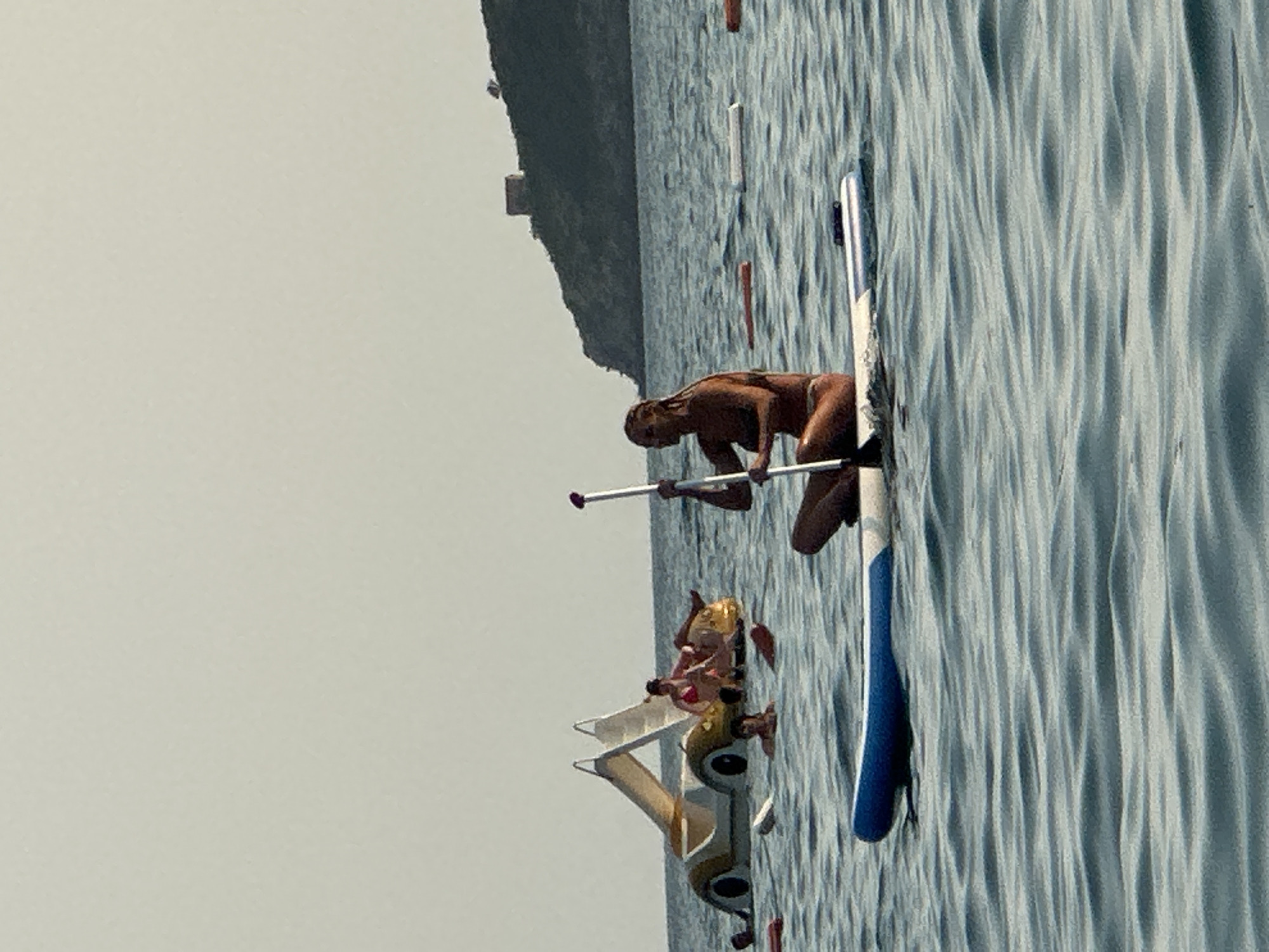 In the city of Boreti, Montenegro, on a clear day, several people are engaged in water activities. In the foreground, a woman with braided hair is paddleboarding, seated on her board and holding a paddle. Her body is turned to the side, and she is looking forward. The paddleboard has a blue stripe along its side. In the middle ground, several people are on a large, novelty watercraft resembling a yellow car with two slides. Two individuals are visible on the "car," with one in a red bikini. Other people are in the water around the craft, some partially submerged. Further in the background, a rocky, tree-covered hill rises from the sea, topped with a small, square stone structure, likely a tower or fortification. The sea is calm with gentle ripples. The sky is a pale, uniform color.