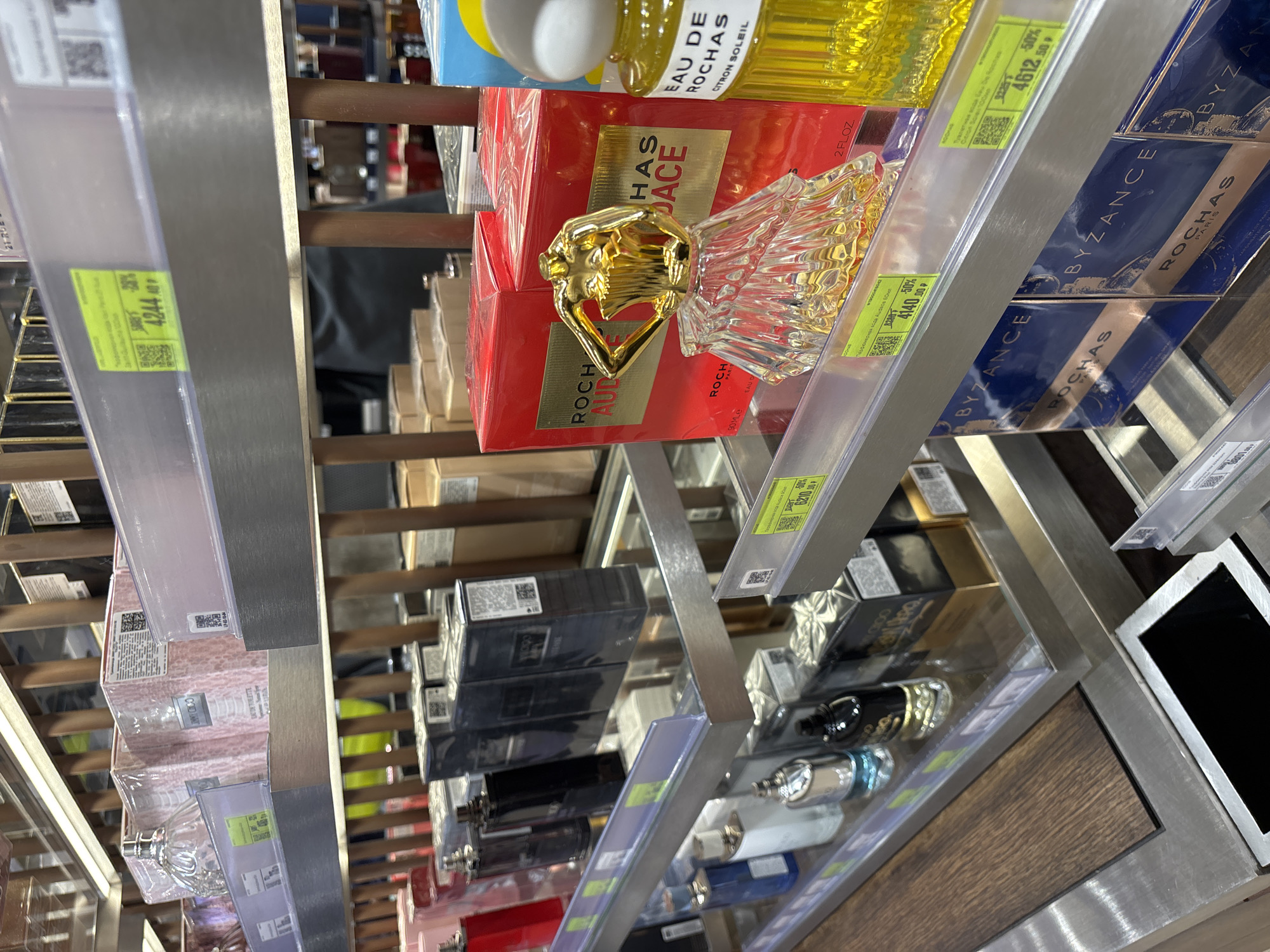 Here's a matter-of-fact description of the image:
**Scene:** The image depicts a shelf of perfume bottles in a store.
**Objects:**
* Multiple perfume bottles are on display. Brands like Rochas are visible. Specific types include "Rochas Audace," "Eau de Rochas," and "Byzance Rochas."
* Price tags with QR codes and Russian text/currency amounts are affixed to the shelf in front of the products.
* Shelving is metal with a clear plastic front piece holding the price tags.
* There is a wood-paneled section visible on the lower right of the frame.
* Part of a digital screen is visible in the lower right of the frame
**Environment:**
* The lighting is bright, likely artificial, as found in a retail environment.
* The focus is on the products on the shelf, indicating a retail setting.
* The presence of price tags suggests the perfumes are for sale.
**Inferred Location (Based on OCR):**
* Given the Russian text and currency amounts on the price tags, the location is likely in Russia. Based on user prompt it is St. Petersburg, Russia.