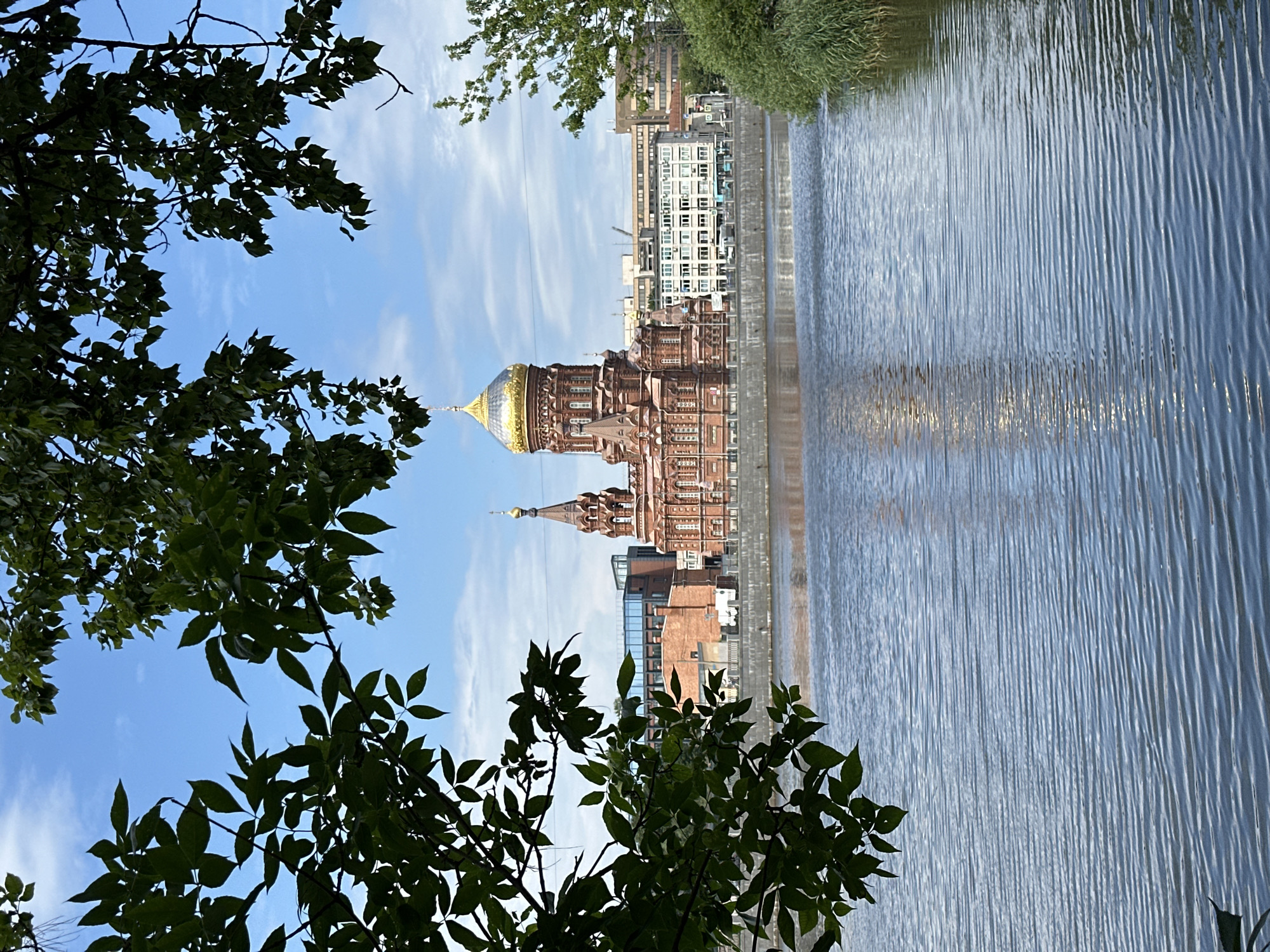 The image captures a view of the Trinity Cathedral, also known as the Smolensky Cathedral, in Saint Petersburg, Russia, from across the Neva River. The cathedral, constructed of red brick with a prominent golden dome and smaller gilded domes, stands as the central subject. It is framed by the lush green foliage of trees in the foreground, which partially obscure the view.
The river itself occupies the lower half of the image, its surface rippled by the wind, reflecting the blue sky and the surrounding buildings. On the opposite bank, beyond the cathedral, a series of modern buildings with white facades and numerous windows are visible, suggesting an urban environment. The sky is a clear blue with scattered white clouds. To the right, a dense patch of green reeds grows along the riverbank. The overall scene is tranquil, depicting a picturesque urban landscape with a prominent religious landmark.