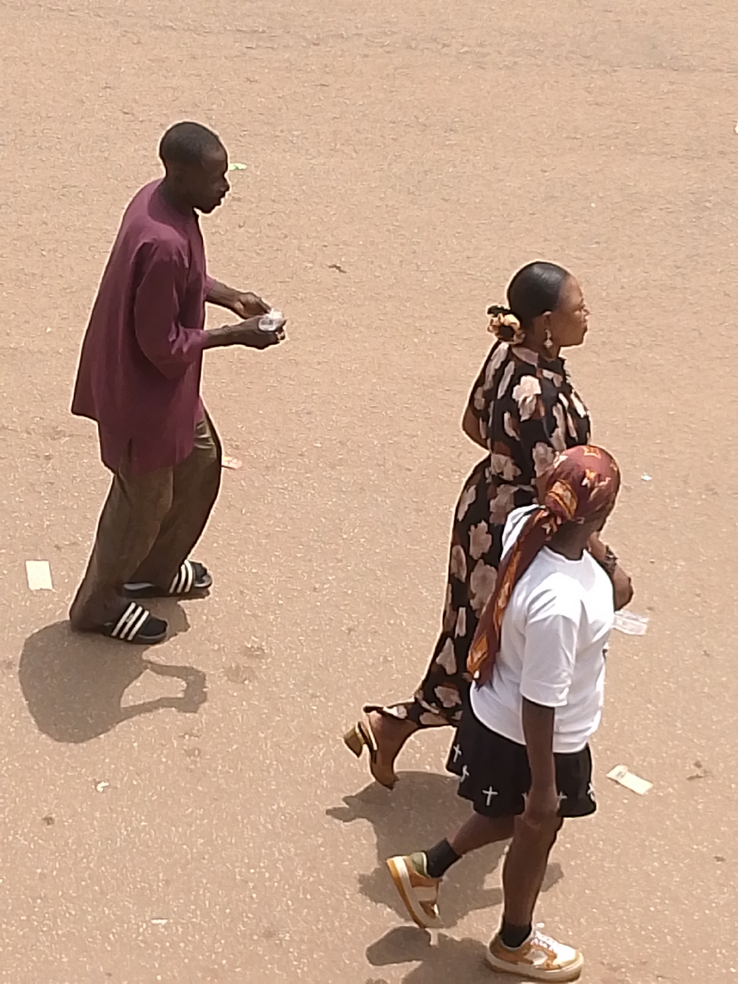Three people are walking on a paved surface in Bukuru, Nigeria. A man is walking in the foreground on the left side wearing a maroon shirt, dark pants, and Adidas-branded sandals. He is holding something in his hands. Slightly behind him, a woman in a patterned long dress and heels is walking in the same direction. To her right, another woman is walking, wearing a white t-shirt, a black skirt with white crosses, and yellow sneakers. She has a reddish-brown headscarf. Pieces of paper or trash are scattered on the ground.