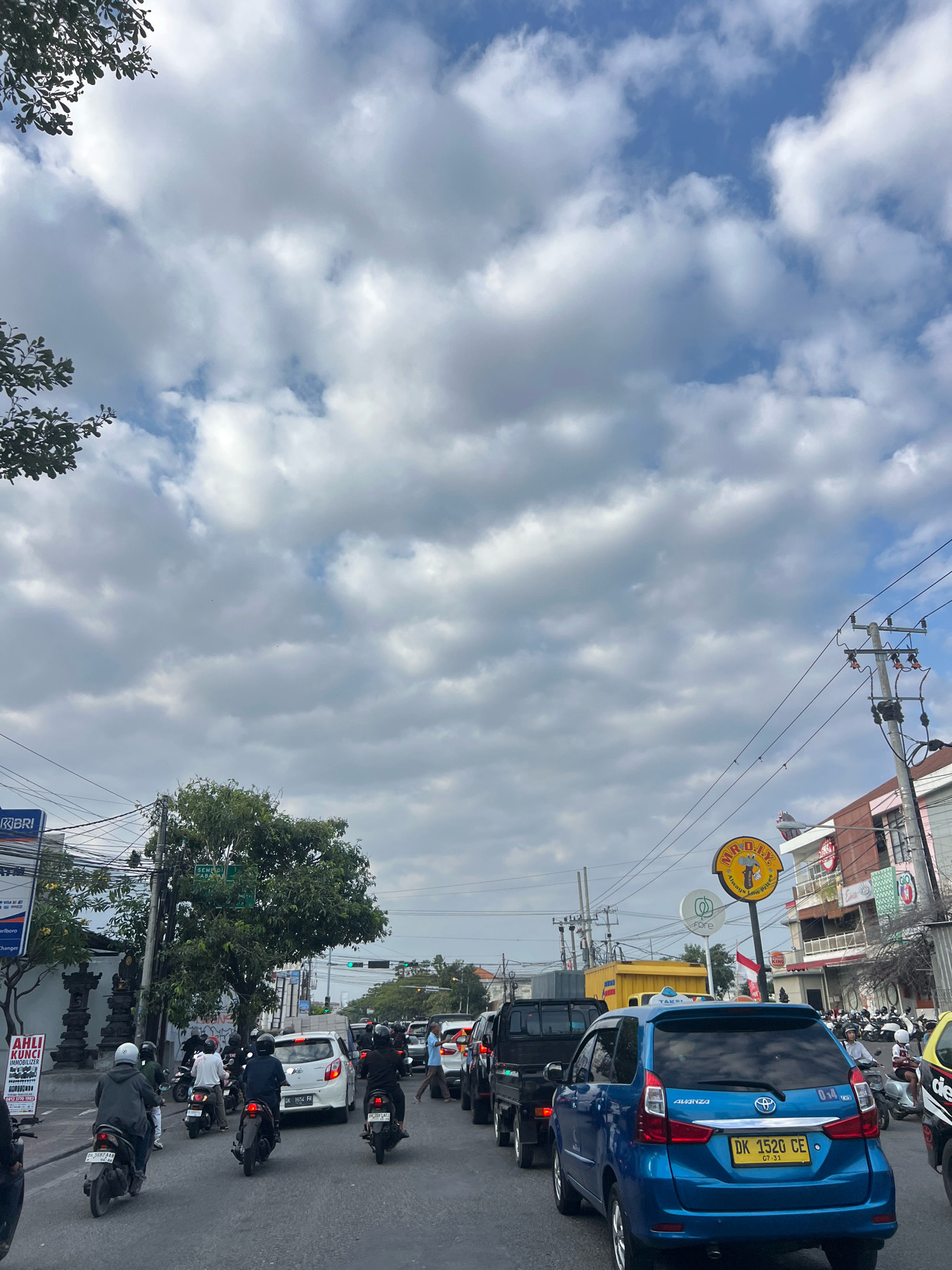 The image captures a congested street scene in Denpasar, Indonesia, under a cloudy sky. The perspective is from within a vehicle, looking forward at traffic.
**Vehicles:** A line of vehicles, including cars, motorcycles, and trucks, fills the road. A blue Toyota Avanza with license plate "DK 1520 CE" is prominent in the foreground. Other vehicles include a white hatchback, a black truck, and numerous motorcycles carrying drivers and passengers, many wearing helmets.
**People:** Several individuals are visible on motorcycles, navigating through the traffic. A person in a blue shirt is standing on the side of the road, to the right.
**Environment:** The street is lined with buildings, some with signage indicating businesses. A large tree provides shade on the left side. Electrical poles and wires crisscross the sky. Road signs are visible, including one with the text "SEMAPUR" and another that appears to be a business advertisement with a circular logo. Traffic lights are visible in the distance, suggesting an intersection. The presence of multiple parked motorcycles indicates a busy area.
**Atmosphere:** The cloudy sky suggests overcast or potentially approaching rain. The traffic jam implies a busy urban environment.
**Location Clues:** The license plate "DK" is specific to Bali, Indonesia, and "Denpasar" is a major city on the island. The architecture of the buildings and the types of vehicles are consistent with an Indonesian setting.