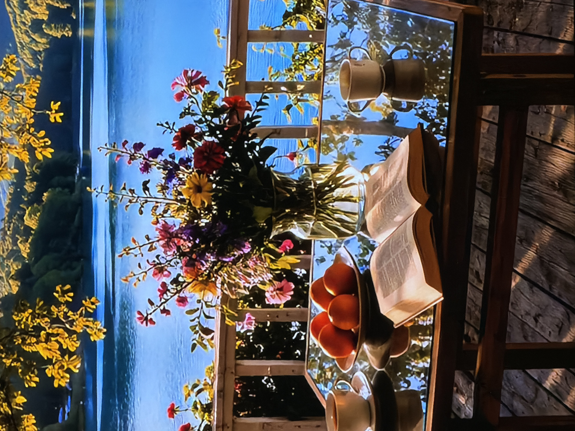 The image depicts a serene outdoor scene, likely on a balcony or patio, overlooking a vast body of water under a clear blue sky. The photo is rotated 90 degrees counter-clockwise from its intended horizontal orientation.
In the foreground, a reflective glass-top table is set with several items. A vibrant bouquet of mixed flowers, featuring various shades of red, yellow, pink, and purple blossoms, stands in a clear glass vase at the center. To the right of the flowers, an open book rests on the table, its pages splayed. Beside the book, a dark bowl is filled with several ripe oranges or tangerines. Further to the right, a light-colored ceramic mug is visible, with a faint reflection of another mug nearby on the shiny table surface. The glass table perfectly mirrors the blue sky, the distant trees, and parts of the wooden railing and surrounding foliage.
The background reveals a tranquil lake or wide river, its calm surface reflecting the bright blue sky above. The far bank is lined with lush green trees, suggesting a forested landscape. In the immediate left foreground, bright yellow-leafed branches frame the view, adding a touch of golden warmth to the natural palette. A wooden lattice or railing forms part of the balcony structure, offering glimpses of more greenery beyond.
The scene conveys a sense of peaceful relaxation, suggesting a quiet moment spent enjoying nature, reading, and a simple meal or drink. The time of day appears to be bright daylight, possibly morning or afternoon, given the strong illumination and clear weather. There are no people visible in the image. No discernible text is visible on the book or any other objects. The provided location context is Sofiivska Borshchahivka, Ukraine, implying this tranquil setting is found within that region.