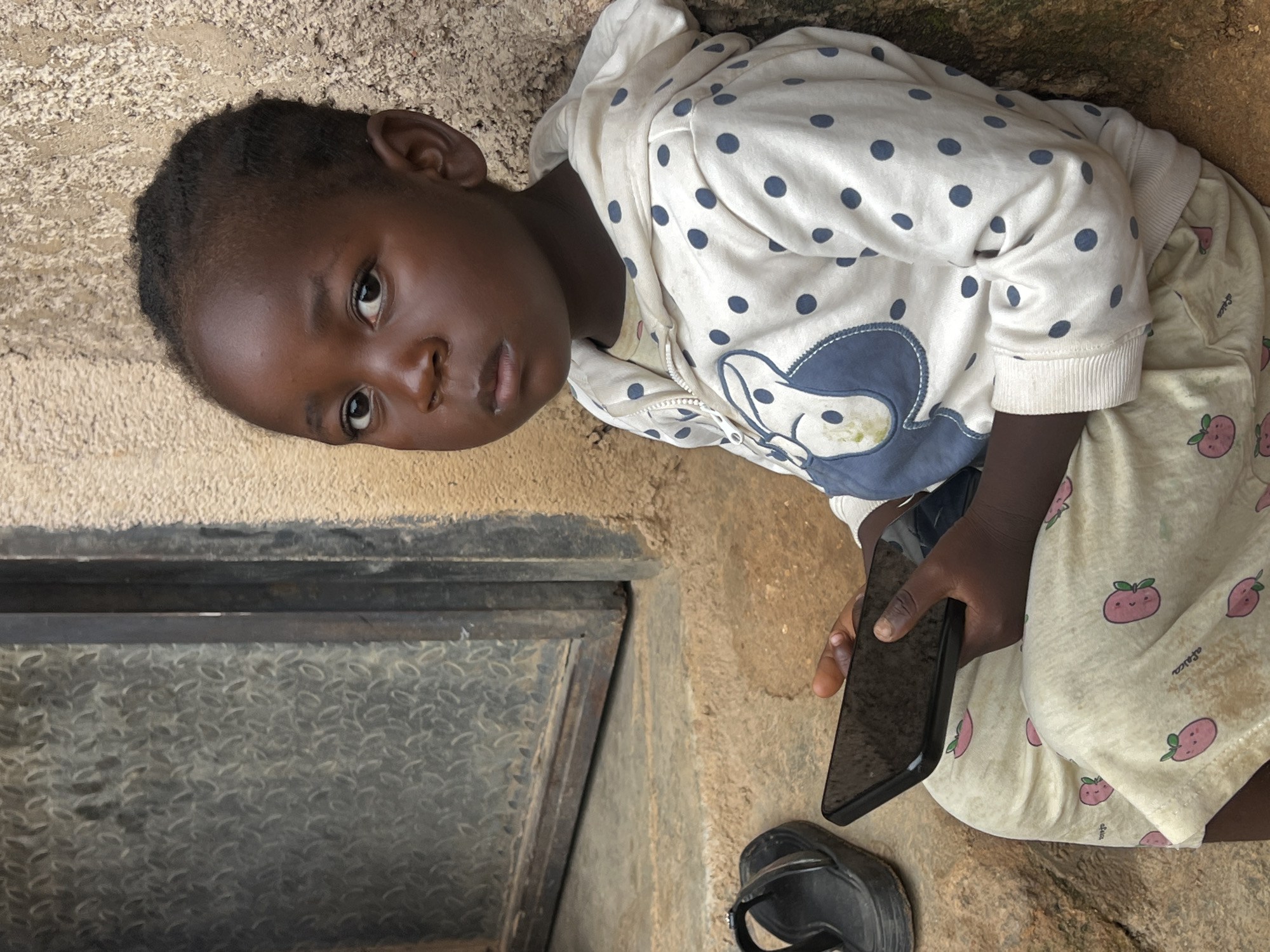 In Jos, Nigeria, a young child is pictured sitting on rough-hewn stone steps next to a building with a textured, beige wall and a metal grate door. The child, wearing a white hooded sweatshirt with navy blue polka dots and a cartoon character on the front, along with pale yellow pants adorned with pink strawberry prints, is holding a black smartphone. A black sandal rests on the step to the left of the child. The scene appears to be outdoors, with the sun casting shadows and highlighting the textures of the environment.