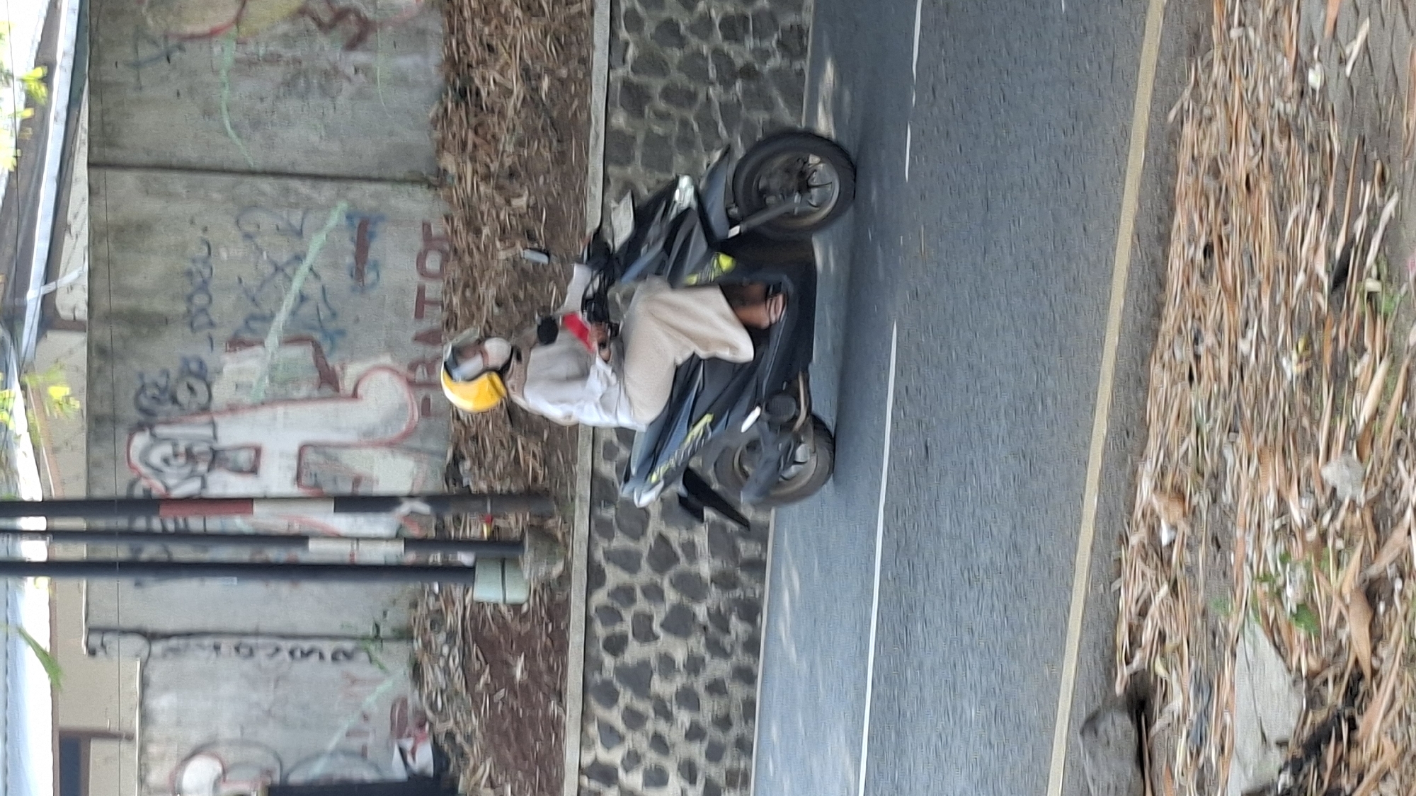 A person wearing a yellow helmet, a light-colored long-sleeved shirt, beige pants, and a face mask rides a black motorcycle on an asphalt road. The motorcycle has a dark body with yellow and black accents. Behind the motorcycle, a stone wall runs horizontally, topped by a concrete wall with graffiti. The ground to the left of the road is covered with dry leaves and debris. The scene appears to be in an outdoor urban environment, possibly in Cilaku, Indonesia, given the context.