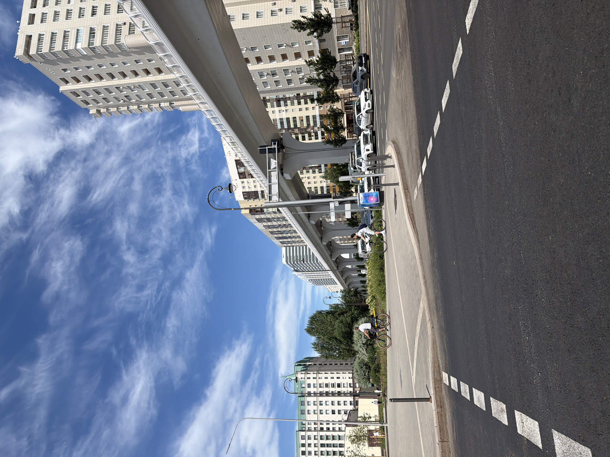 This is a wide-angle, vertical shot of an urban scene in Astana, Kazakhstan. The image is dominated by a modern residential building and an elevated monorail track that runs along its side. The sky is a vibrant blue with wispy white clouds, suggesting a clear and pleasant day.
In the foreground, a paved road with white dashed markings runs parallel to the sidewalk and a grassy area. A few cars are parked along the roadside. Further into the scene, two individuals are seen riding bicycles on the sidewalk. One cyclist is dressed in a white t-shirt and light-colored shorts, while the other is in a yellow shirt and dark shorts. They appear to be enjoying a leisurely ride.
Behind the cyclists, there is lush greenery and more residential buildings, some with distinct architectural features like green roofs and balconies. The overall impression is one of a modern, well-developed city with ample green spaces and amenities for its residents. The lighting suggests it's daytime, possibly late morning or early afternoon, with the sun casting shadows and illuminating the buildings and road. The scene evokes a sense of calm and everyday life. No visible text is present in the image.