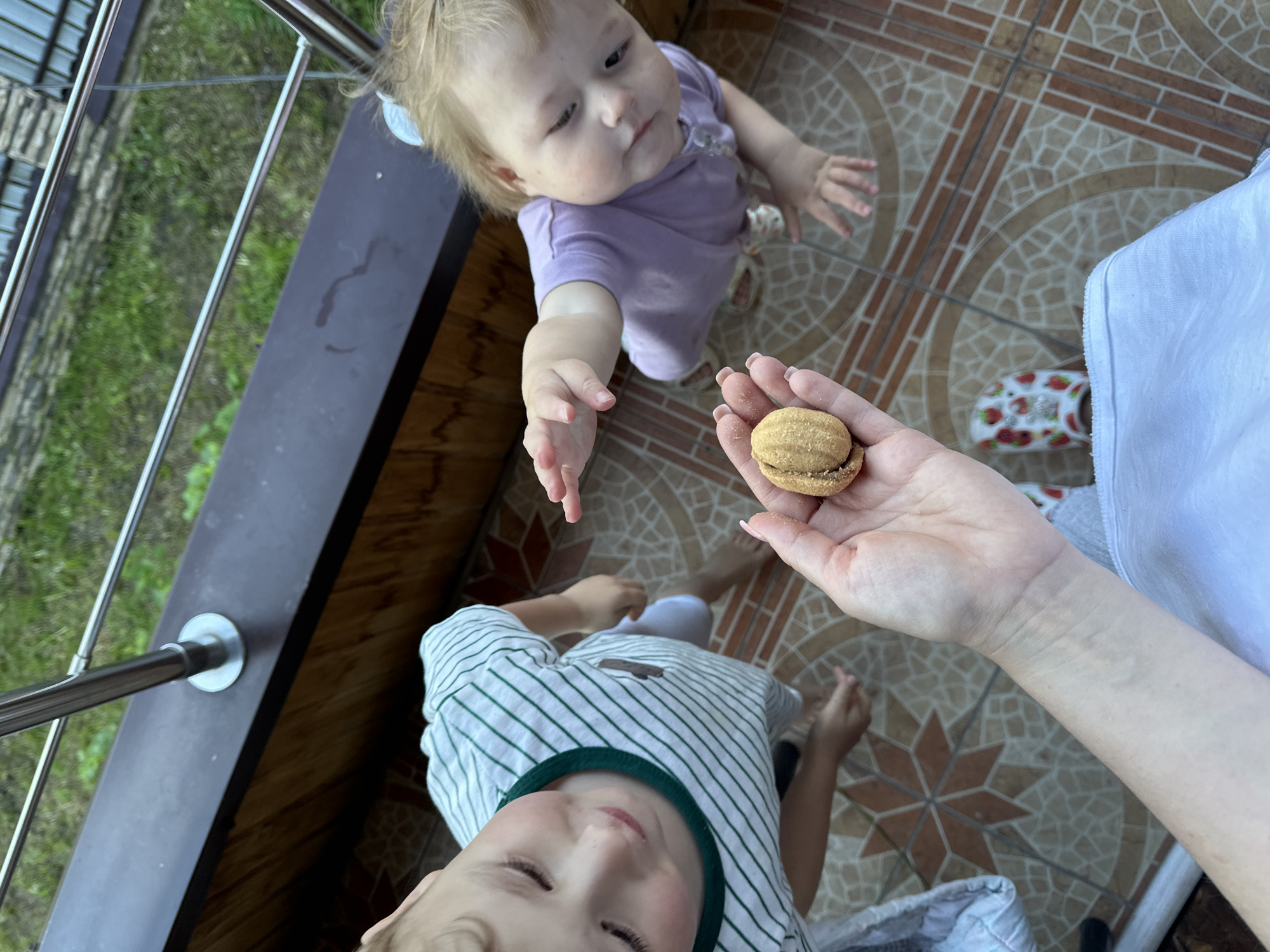 A top-down shot features two children and an adult on a tiled balcony. The adult holds a walnut-shaped cookie in their hand, while a younger child in a purple shirt reaches for it. A slightly older child in a striped shirt looks upwards, seemingly towards the cookie. The tiled balcony floor has a brown and beige geometric pattern. A railing runs along the edge of the balcony, with greenery visible beyond. One sandal with a strawberry print is also visible. The location is Nizhny Novgorod, Russia.