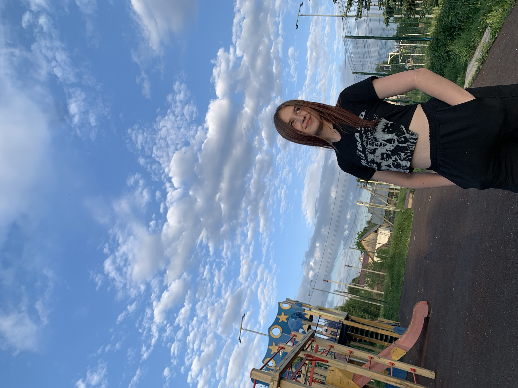 The image depicts a young woman standing in a park. She is wearing a black crop top and black shorts. The background shows a playground structure with slides and climbing elements. Beyond the playground, there's a fenced-in sports area, possibly a basketball or tennis court. The sky is mostly blue with numerous cumulus clouds. There are houses visible in the distance, suggesting the park is located in a residential area. The park area is paved with asphalt. Street lights are present. It can be determined that the location is Фоки, Russia based on information about the location of the photo.