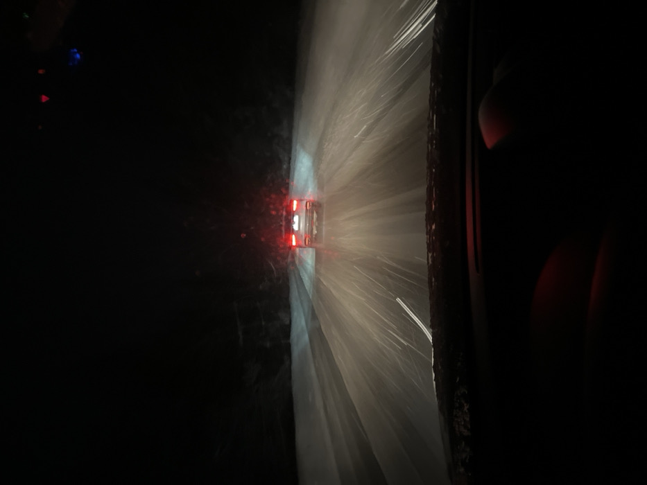 The image captures a nighttime scene on a snow-covered road in Arbuzovo, Russia, viewed from the perspective of a driver inside a vehicle. Heavy snowfall is actively illuminated by vehicle lights. A light-colored sedan is positioned ahead in the same lane, its red taillights illuminated. Headlights from the camera's vehicle cast prominent beams forward, intensely illuminating the falling snow particles which appear as bright streaks, and reflecting off the snow-covered road surface. The road itself is uniformly covered in snow, with faint disturbances visible where vehicles have passed. On both sides of the road, dark, indistinct masses suggest dense, snow-covered trees or vegetation under a completely dark sky. The lower portion of the image reveals the top of a vehicle dashboard and a human hand, possibly a right hand, resting on the dashboard area on the lower right, faintly illuminated with a reddish glow from inside the vehicle. Visibility is significantly impaired by the darkness and heavy snowfall.