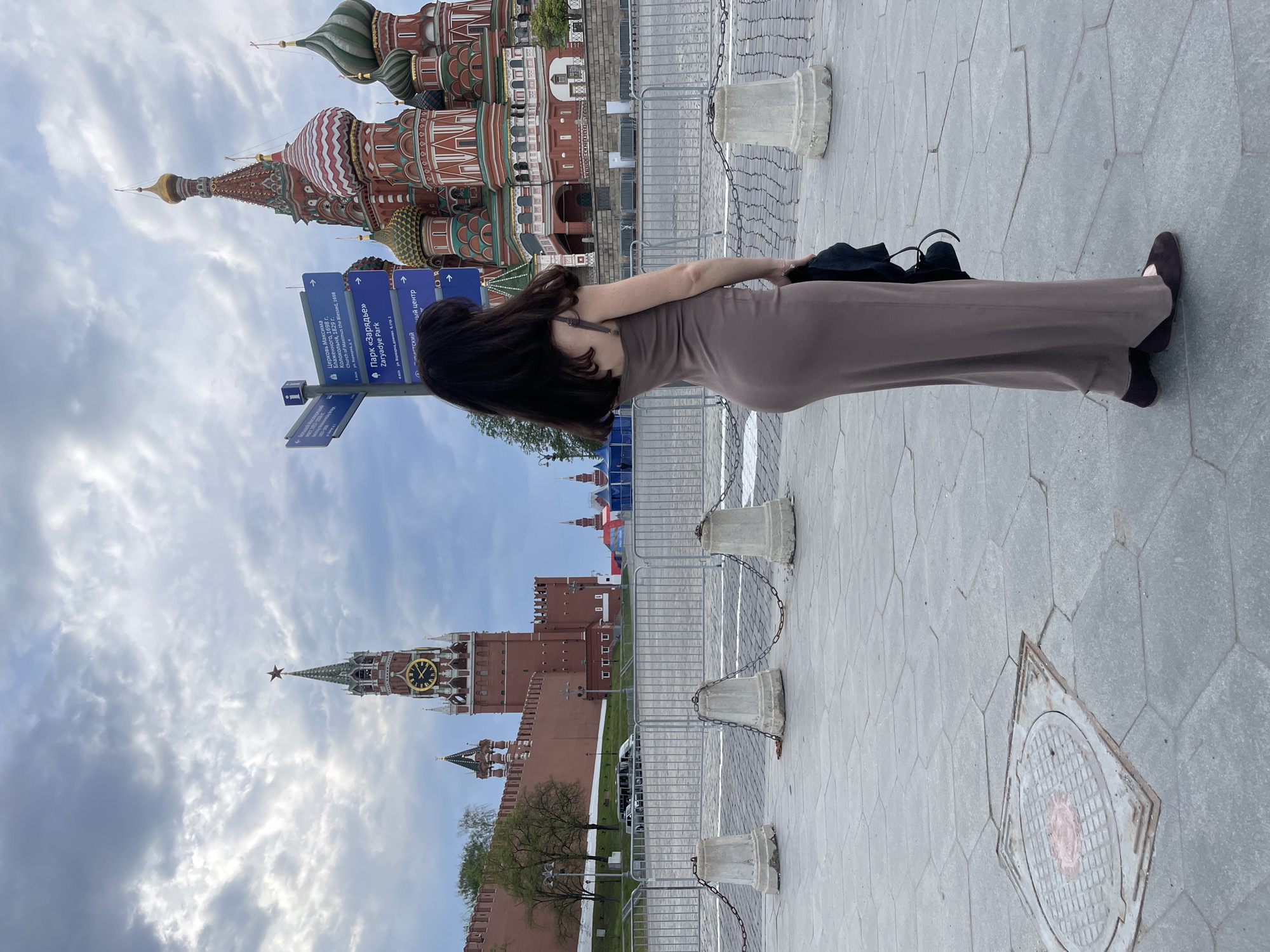 The image shows a woman standing on a gray stone paved area, possibly a square, with her back towards the camera. She is wearing a long, fitted brown dress and black shoes. Her long dark hair hangs down her back. In the background, there are several distinct buildings, including the colorful and ornate St. Basil's Cathedral with its onion-shaped domes, as well as a red brick building with a clock tower, likely part of the Moscow Kremlin. Blue street signs with white lettering are visible nearby. Concrete barriers connected by chains line the edge of the walkway. The sky is partly cloudy. The location is likely Red Square in Moscow, Russia.