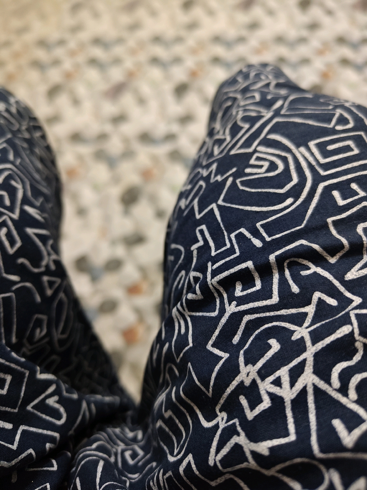 The image shows a close-up view of two dark blue pants with a white abstract geometric pattern. The pants are draped, suggesting a person is wearing them. The background is out of focus and appears to be a speckled floor or surface with a light tan and cream color palette, possibly tiles or terrazzo. The lighting is soft and diffused. The image does not provide any information about the city of Garhwa, India.