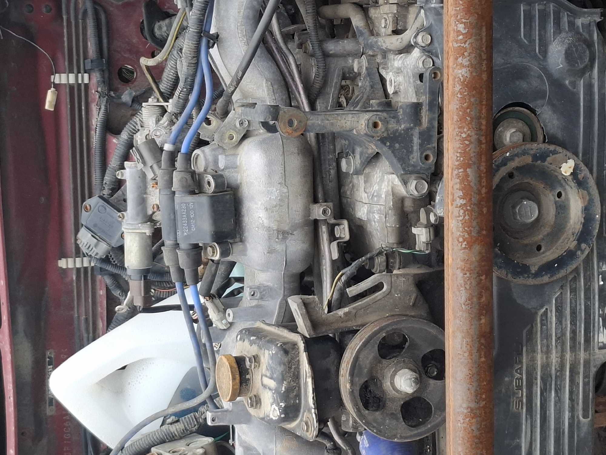 The image is a close-up, eye-level, outdoor photograph of a car engine, likely taken in Almaty, Kazakhstan, though there are no specific visual indicators of this location. The engine is dirty and appears to be from an older vehicle, possibly a Subaru, as indicated by the "SUBARU" text on a black engine component at the bottom of the frame.
The engine is a complex assembly of metal parts, wires, and hoses. Prominently visible are:
* **Engine Block and Components:** A large, metallic engine block is central to the image. Various aluminum and cast iron parts, including intake manifolds and covers, are visible. These are coated in dust and grime.
* **Fluid Reservoir:** A white plastic fluid reservoir with a partially filled, foamy liquid is located in the upper left quadrant of the image.
* **Wiring Harness:** Numerous bundles of electrical wires, insulated with black plastic and some with blue sleeves, are routed throughout the engine bay. Some are secured with zip ties.
* **Pulleys and Belts:** A large, dark-colored pulley with three circular cutouts is visible on the left side of the image, connected to the engine by a shaft. A belt likely runs over this pulley.
* **Sensors and Connectors:** Various electrical sensors and connectors, typically gray or black plastic, are attached to different parts of the engine.
* **Fasteners:** Numerous bolts and nuts are visible, securing various engine components.
* **Corrosion:** A thick, rusty metal pipe or bar runs horizontally across the lower middle of the image, obscuring some of the engine details. This suggests potential exposure to the elements or the age of the vehicle.
* **Other Parts:** A cylindrical gold-colored cap is on top of a black component, likely an oil or fluid cap.
The lighting is relatively even, suggesting it is daytime. The background is out of focus but appears to be the engine bay of a vehicle. There are no people, animals, or specific landmarks visible in the image. The overall impression is of a functional but neglected automotive engine.