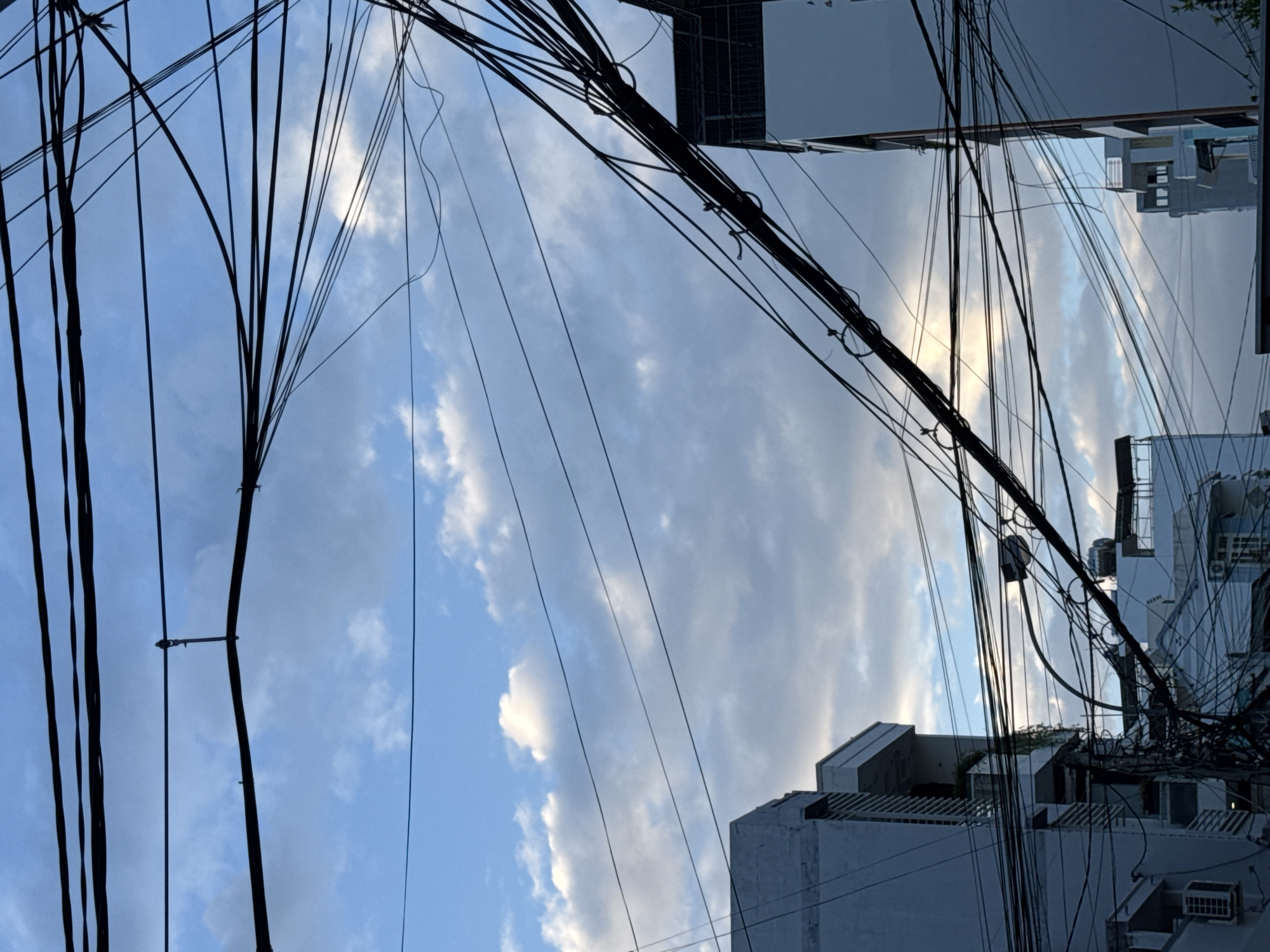 The image depicts a dense network of overhead electrical wires against a cloudy blue sky. Several modern buildings with white facades and balconies are visible in the lower portion of the frame, suggesting an urban setting. The composition is dominated by the intricate web of wires, which crisscross the sky and obscure portions of the buildings. There are no people visible in the image. The scene is characteristic of areas in Nha Trang, Vietnam, where such extensive overhead wiring is common.