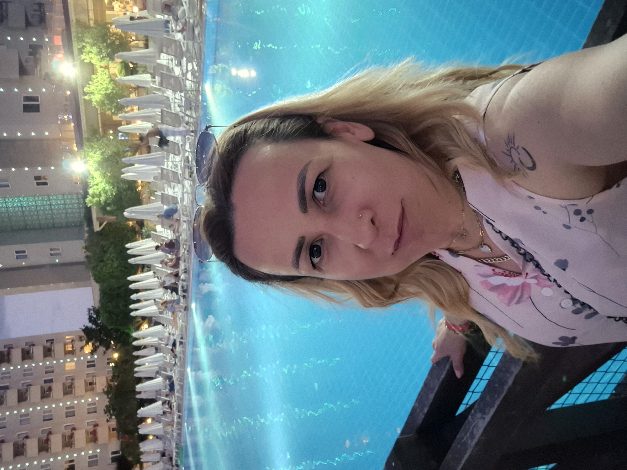 A woman with blonde hair and sunglasses on her head is taking a selfie in front of a swimming pool at dusk. She is wearing a light-colored blouse with a floral pattern. In the background, there are lounge chairs and umbrellas arranged around the pool, as well as a multi-story building. The pool water is illuminated by underwater lights. The scene appears to be at a resort or hotel.