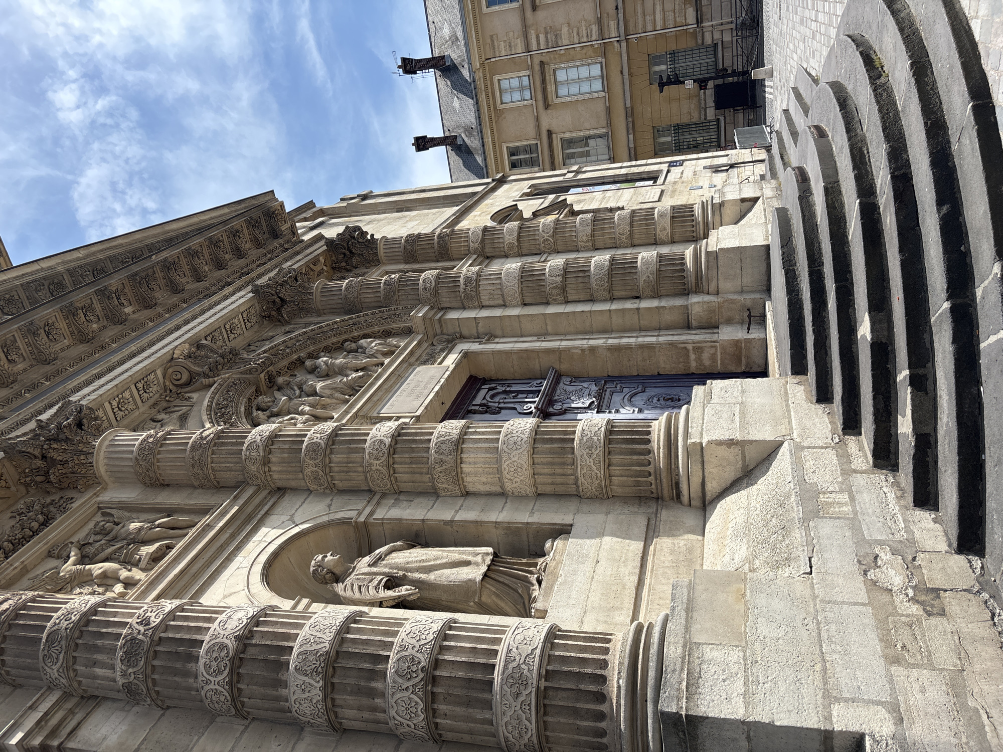 This is a low-angle, wide shot of the ornate facade of a historical building in Paris, France. The image is oriented vertically, giving a dramatic upward perspective. The architecture is characterized by intricate stone carvings, prominent fluted columns, and decorative friezes.
The building features multiple levels with large, arched windows and balconies. The stonework is weathered, suggesting age and historical significance. On the left side, a series of detailed sculptures depict human figures, possibly mythological or religious in nature, integrated into the architectural design. Niches within the facade hold statues, one of which appears to be a robed figure. The building's surface is a light-colored stone, contrasting with darker, possibly stone, arched elements visible on the far right, which seem to be part of an entrance or a grand staircase.
Above the main facade, other parts of the building are visible, including a steeply pitched roof with chimneys and upper stories with smaller windows. The sky above is a bright blue with wispy white clouds, indicating a clear, sunny day, likely mid-morning or early afternoon due to the strong, directional lighting that casts shadows across the architectural details.
The scene conveys a sense of grandeur and historical depth, typical of Parisian architecture. There is no discernible activity or event taking place; the focus is solely on the architectural splendor of the building. No visible text is present in the image.