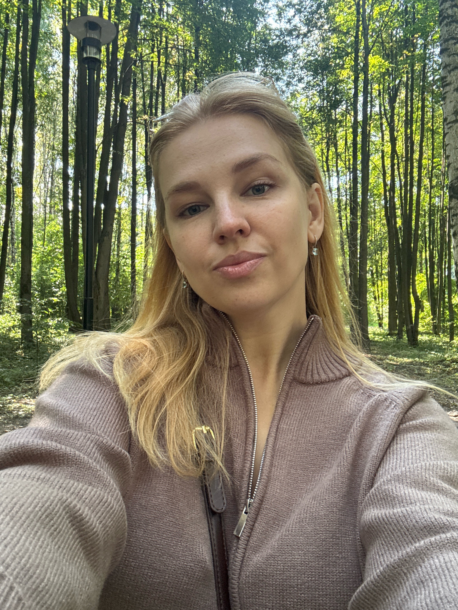 A young woman with blonde hair is taking a selfie outdoors. She is wearing a zipped-up, light brown knitted sweater. She has light blue eyes and is wearing small, square earrings. Behind her, a line of tall trees with green leaves creates a natural backdrop. Sunlight filters through the trees, illuminating the scene. To the left of the woman, a black lamppost with a round, metallic light fixture is visible. The ground appears to be covered with dry leaves and dirt, suggesting a park or forest setting. The image was taken in Rusanovo, Russia.