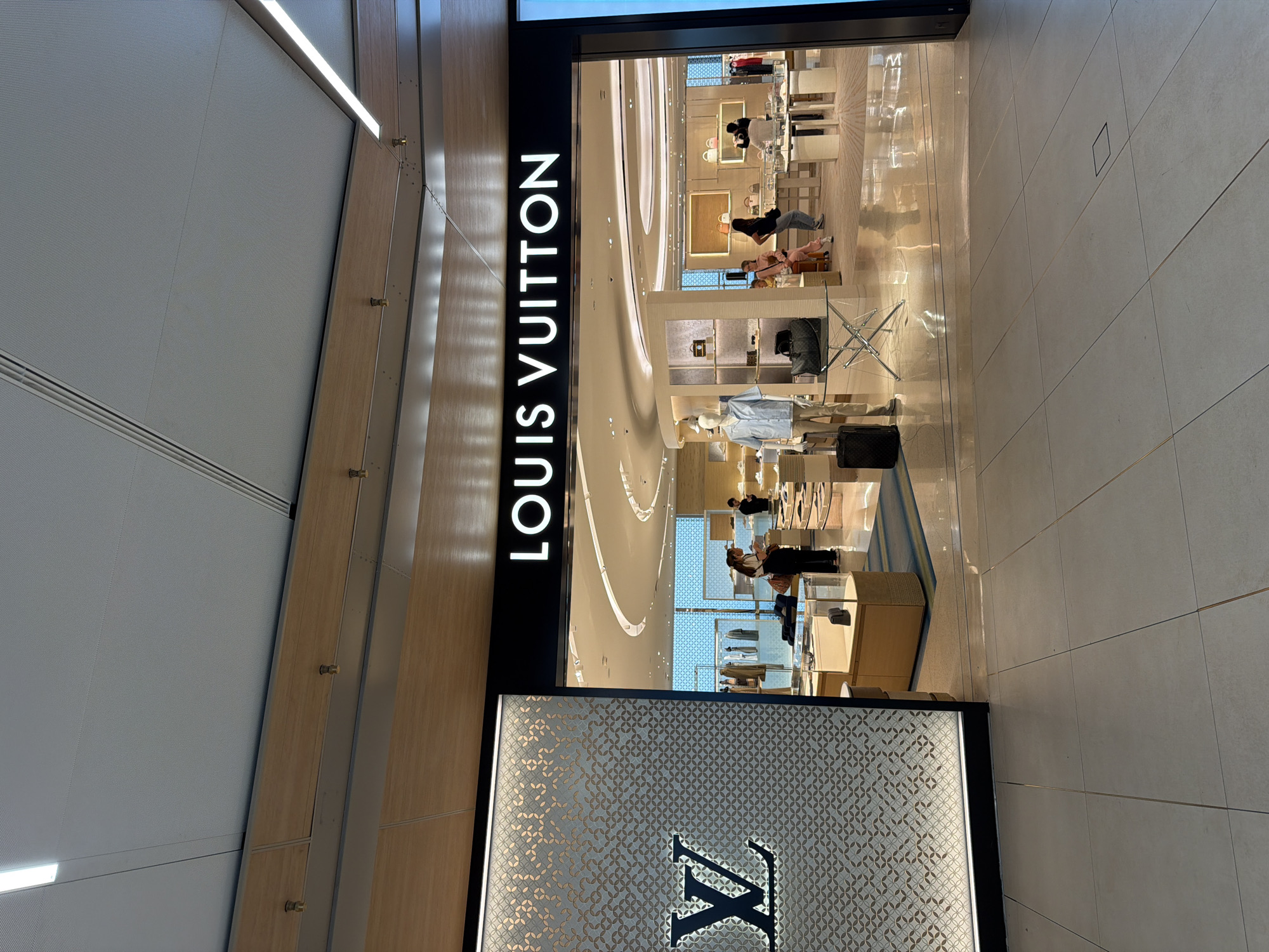 The image shows the entrance to a Louis Vuitton store. The store's name is displayed in large white letters on a black background above the entrance. To the left of the entrance is a wall decorated with a patterned mosaic design and a large Louis Vuitton logo. Inside the store, several display cases with merchandise are visible, as well as mannequins dressed in clothing. A few people are browsing in the store. The floor is tiled. The overall environment appears to be a well-lit, modern retail space, likely a luxury boutique.