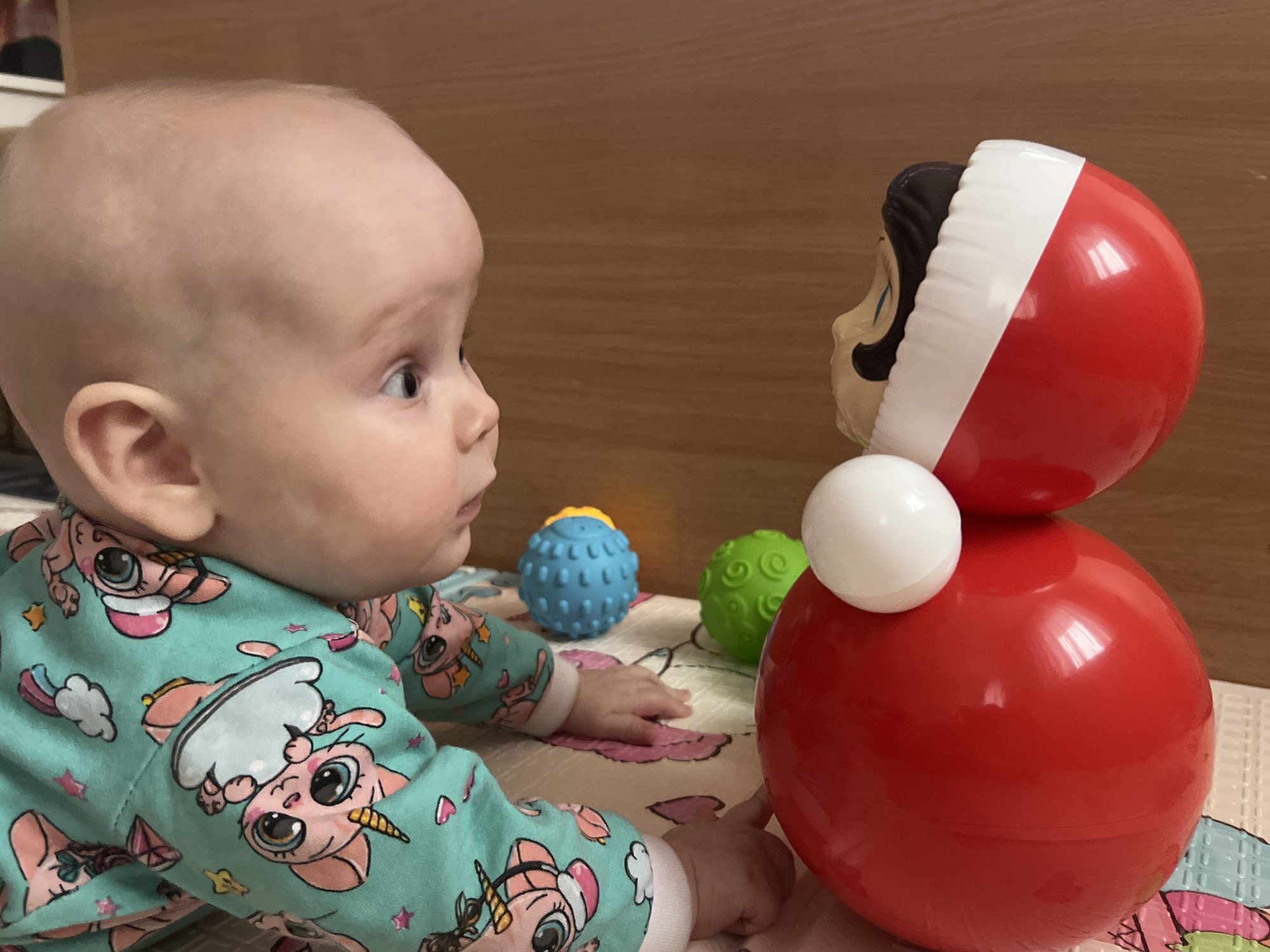 A baby wearing turquoise pajamas with pink and white cartoon unicorns lies on a patterned blanket, looking at a red and white toy with a face. Two small, textured balls, one blue and one green, are nearby on the blanket. The background is a wooden surface. The location and city of Votkinsk, Russia, are not discernible from the image.