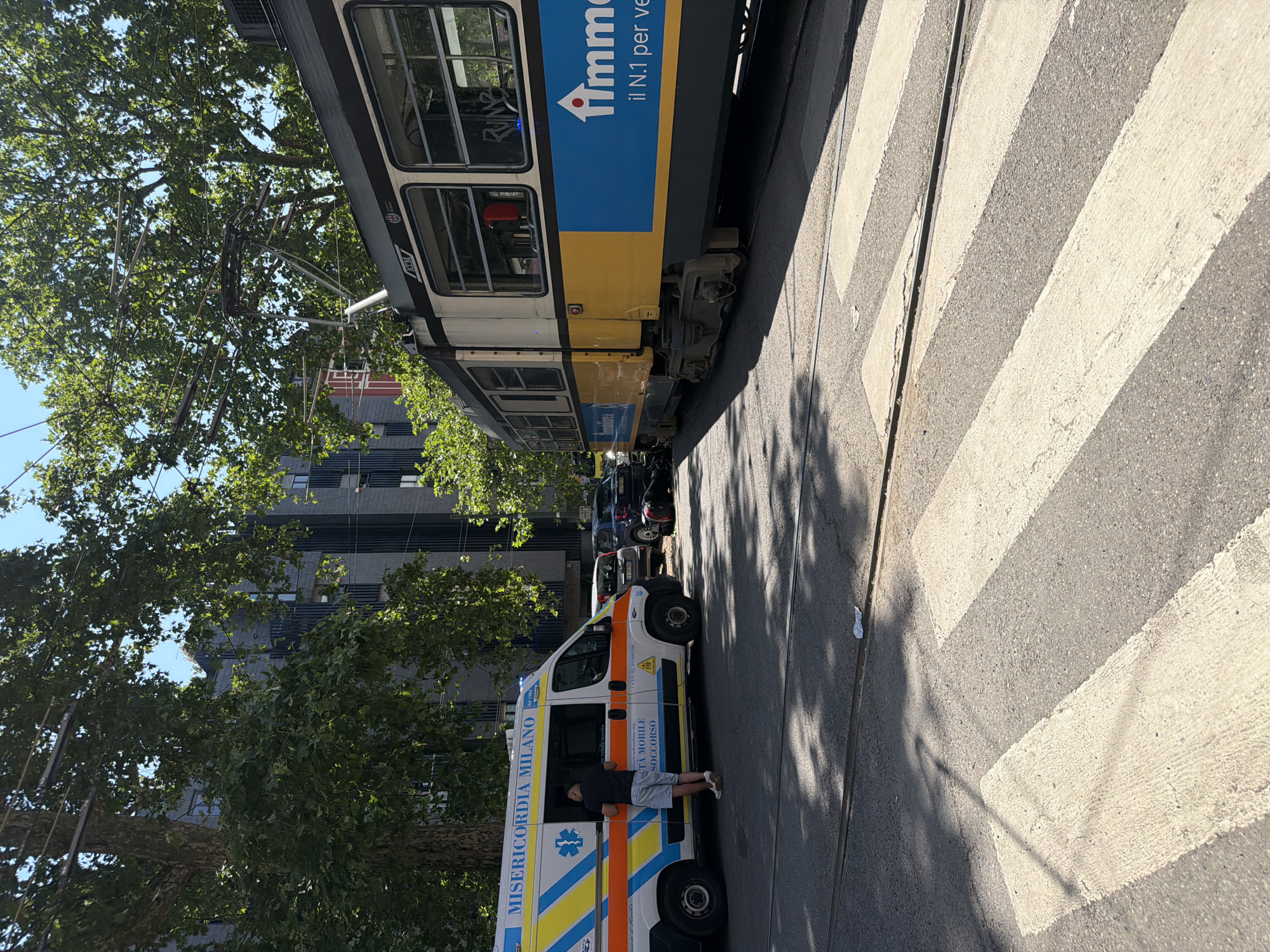 A brightly lit scene in Milan, Italy, shows an ambulance parked near a tram line at a crosswalk. The ambulance, labeled "MISERICORDIA MILANO," with Italian text, is white with orange, blue, and yellow stripes. A person stands at the open door. A yellow and white ATM tram runs along the tracks parallel to the crosswalk. Trees line the street, casting shadows. A multi-story building stands in the background. Several parked cars are seen between the ambulance and the tram.