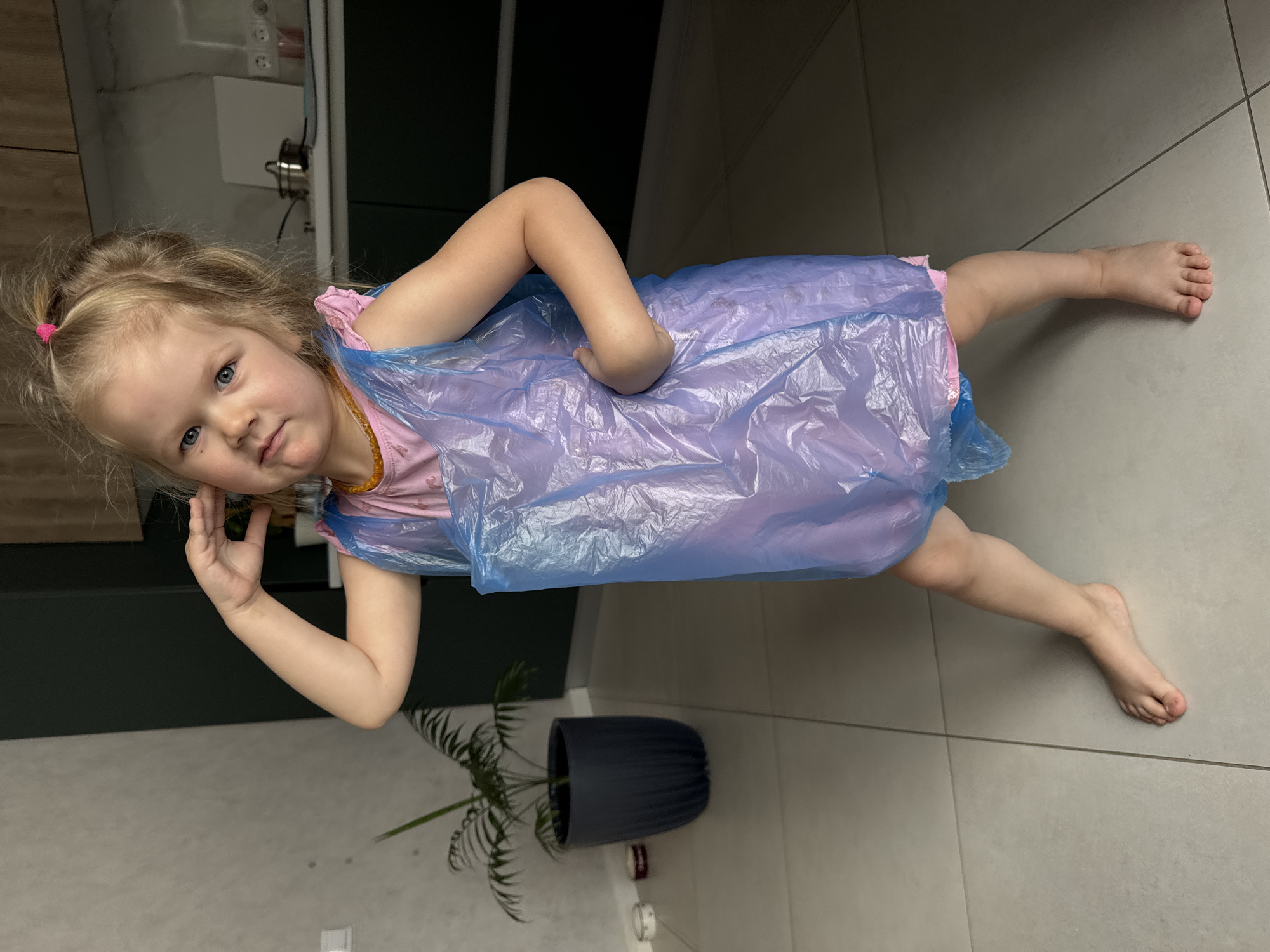 A young girl is standing on a tiled floor in what appears to be a kitchen or dining area. She has blonde hair styled with a pink hair tie, and she is wearing a pink shirt with an amber necklace. Over her shirt, she is wearing a light blue plastic bag as a makeshift dress. She has her left hand raised towards her face. Behind her and to the left is a dark green cabinet and a potted plant with green leaves. The floor is covered with large gray tiles, and the background shows light wood cabinets and countertops, possibly in a kitchen setting. The location is Gornyi (Dvurechenskoe S/P), Russia.