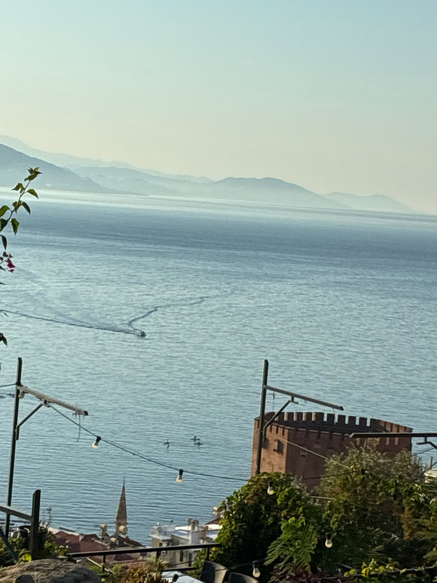 The image depicts a scenic view overlooking the Mediterranean Sea in Alanya, Turkey. In the foreground, a portion of a brown, crenellated tower, likely part of Alanya Castle, is visible on the right side. Lush green foliage and some outdoor lighting fixtures are also present in the foreground. The middle ground features the tranquil blue waters of the sea, with a small boat leaving a wake as it travels across the surface. In the distance, a series of layered mountains fade into a hazy sky, characteristic of the coastal landscape. The overall scene is bathed in soft, natural light, suggesting either early morning or late afternoon. There are no people clearly visible in the image.