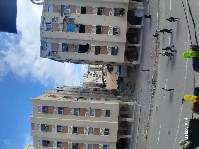 The image depicts an urban residential street scene in Khan Yunis, Palestinian Territory, under a partly cloudy blue sky. The perspective is elevated, with a partial view of colorful clothespins on an unseen foreground structure and "Galaxy A14" text visible in the lower left. A dark, irregular object partially obstructs the top right of the frame.
A paved road with faded white markings runs across the midground. Approximately ten individuals are present: a group of six to seven males are positioned in the street, with one individual prominently wearing a blue top. Another male stands individually on the left side of the street, near the sidewalk. On the right sidewalk, a lone female figure in a dark garment walks away from the viewer, while another individual is seated further along the same sidewalk. On the left sidewalk, a single, smaller individual stands alone near the curb, facing the buildings.
Two prominent multi-story residential buildings, each approximately six to seven stories tall, frame a rubble-strewn gap in the center. Both buildings feature light tan facades with brown horizontal bands and decorative white patterns around windows. The building on the right exhibits extensive damage, including numerous impact marks or bullet holes across its facade, shattered windows, and some openings covered with dark material or a blue tarp. Exposed structural elements are visible in damaged areas. The building on the left also shows signs of damage, with impact marks and broken windows, though appearing less severe than the right structure. Both buildings feature arched entrances or porticoes on their ground floors. The unpaved area between these main buildings is covered in debris, sand, and rubble. In the background, beyond this debris field, additional multi-story residential buildings are visible. The overall scene indicates significant damage to the urban residential infrastructure.