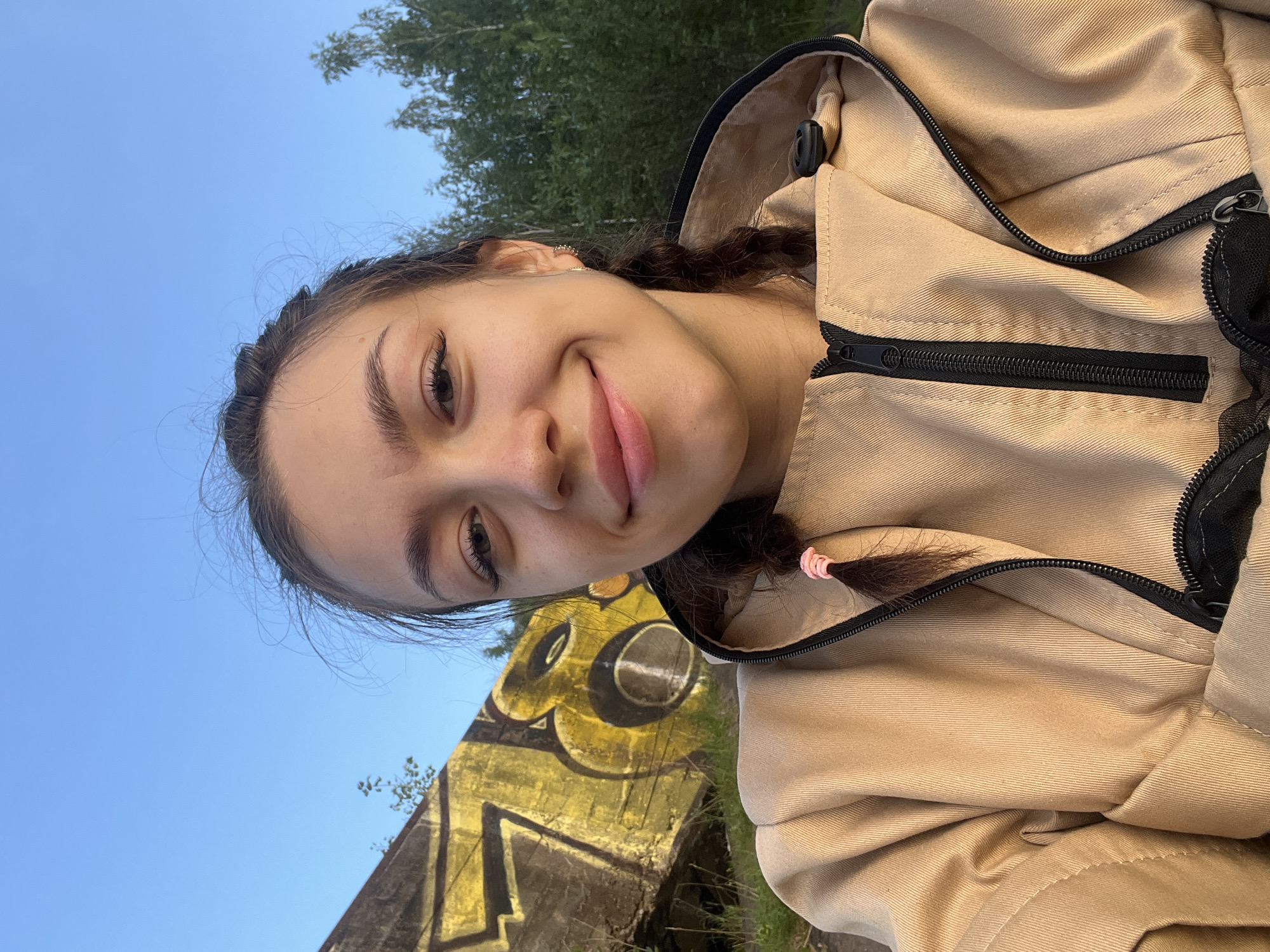 The media file is a selfie taken by a young woman with dark hair styled in two braids, one of which is tied with a pink elastic band. She is wearing a light brown hooded jacket with a black zipper. Her expression is a gentle smile, and her eyes are looking towards the camera.
The background of the image shows a bright blue sky with some trees visible at the top. In the lower part of the frame, there is a concrete structure covered in yellow and black graffiti. The lighting suggests it is daytime, possibly late afternoon or early evening, due to the warm tones. The overall setting appears to be outdoors, likely in a rural or semi-urban area, with evidence of an abandoned or neglected structure. The graffiti on the wall contains stylized lettering and circular shapes. No discernible text is visible in the media file. The media file appears to be taken in Chernushka, Russia.