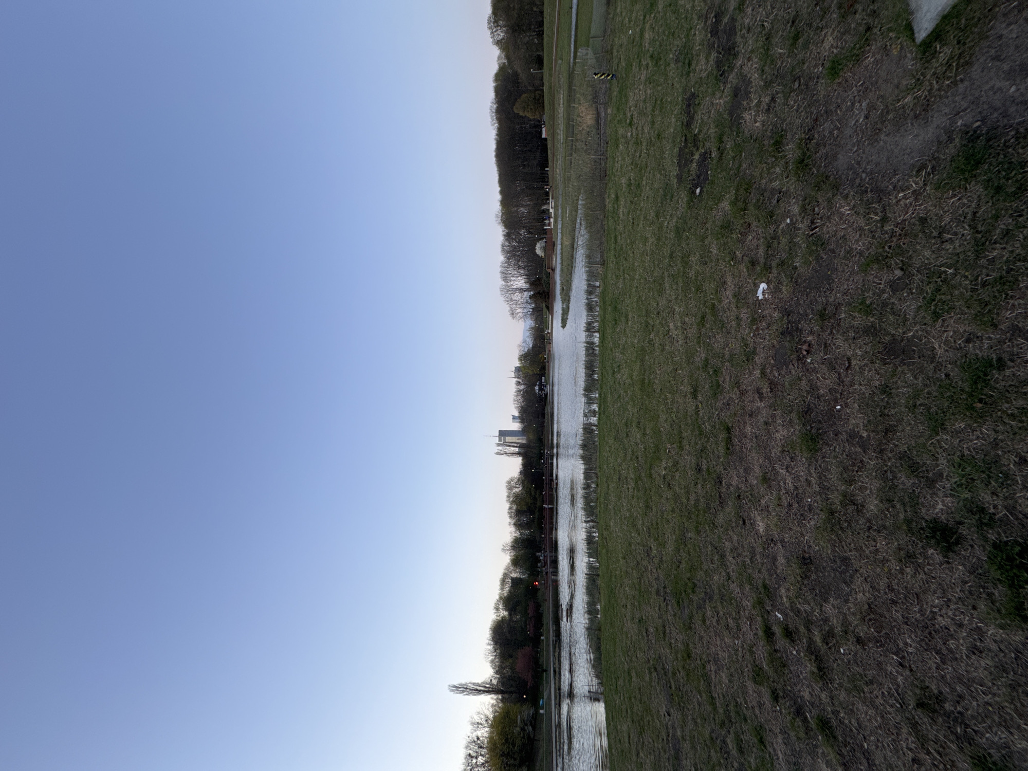 The image depicts a landscape scene in Warsaw, Poland, showcasing a park or open space adjacent to a body of water. The composition is divided into thirds with a large portion of sky that transitions from light blue at the top to a slightly darker hue near the horizon. The middle section features a body of water reflecting the sky and surrounding landscape, lined by trees and vegetation. In the distance, hints of the Warsaw skyline are visible. The foreground and right side of the image consist of a grassy area, with some patches of dry or less vibrant vegetation. The perspective is from ground level, looking across the landscape.