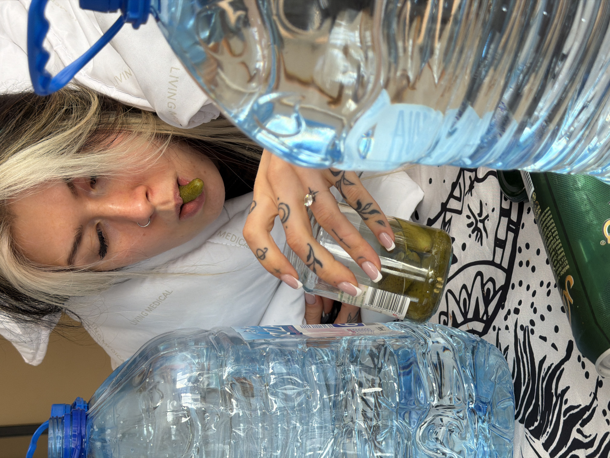 The image depicts a person with light and dark blonde hair lying on a white pillow with the text "LIVING MEDICAL" printed on it. The person has a pickle in their mouth, and their right hand holds a jar of pickles. The person is wearing a ring on their ring finger. Two large bottles of water are positioned above and below the person's face, partially obscuring the view. Next to the pickles, there is a green can with text visible. The background consists of a black and white patterned cloth. The location or city cannot be determined from the provided image details.