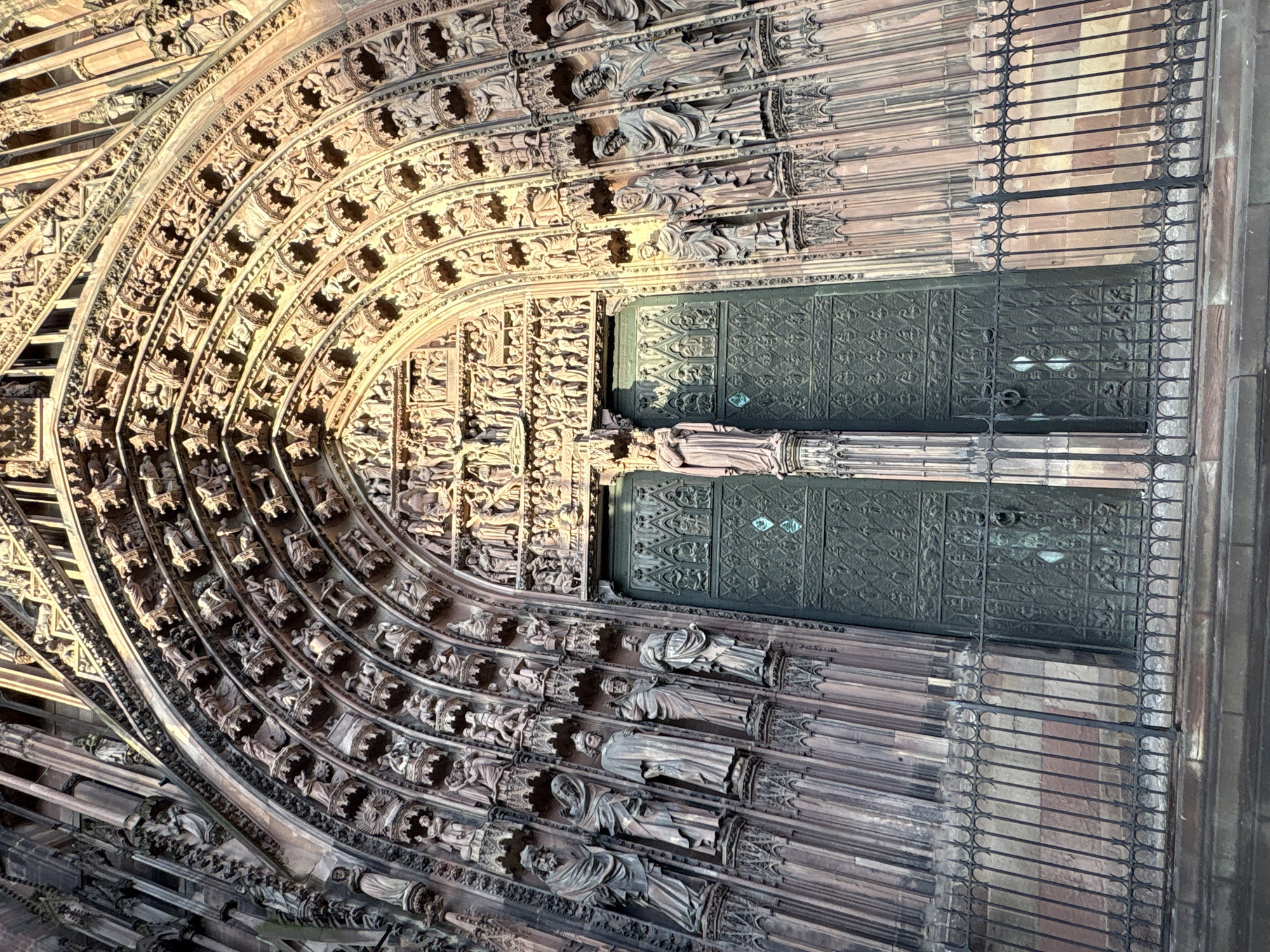 This image captures an exterior view of the Strasbourg Cathedral in France, specifically focusing on the grand entrance. The ornate architectural details include a large, dark, intricately decorated door framed by an arched opening. The archway is adorned with layers of carved figures and decorative motifs. To the right of the door, a metal barrier or gate is in place, likely for protection or crowd control. The stonework is predominantly brown, with variations in tone and texture that suggest age and weathering. The lighting casts shadows across the carvings, enhancing their depth and detail. There are no visible people within the immediate frame of the entrance.