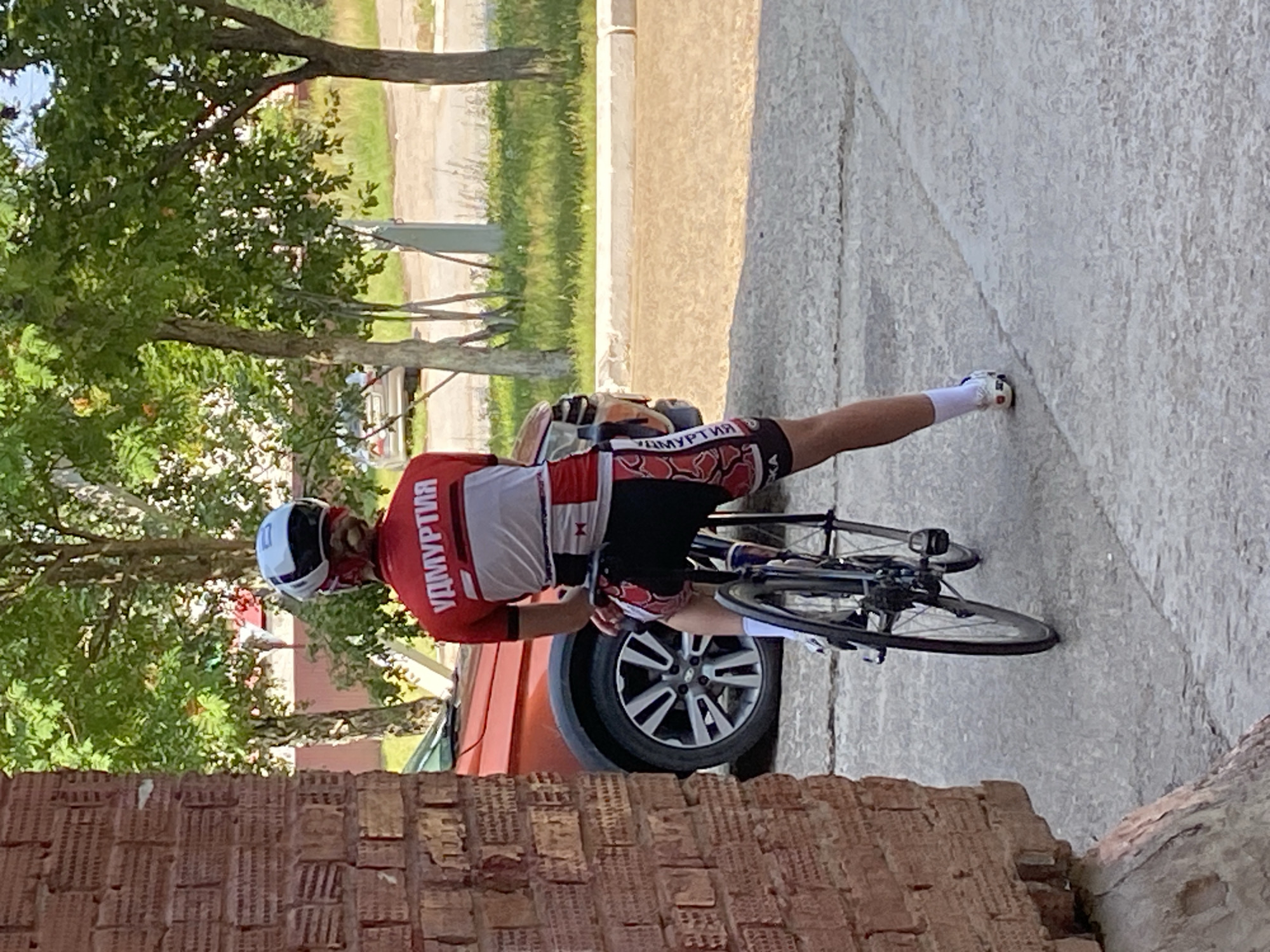 The image depicts a person wearing a cycling helmet and a red and white cycling outfit, with "УДМУРТИЯ" visible on the back of the jersey. They are sitting on a bicycle with black wheels and frame, facing away from the camera. The background shows trees, grass, and a red car parked next to the cyclist. A brick building is visible to the left of the frame. The scene appears to be outdoors on a paved surface. The location is identified as Noviy, Russia.