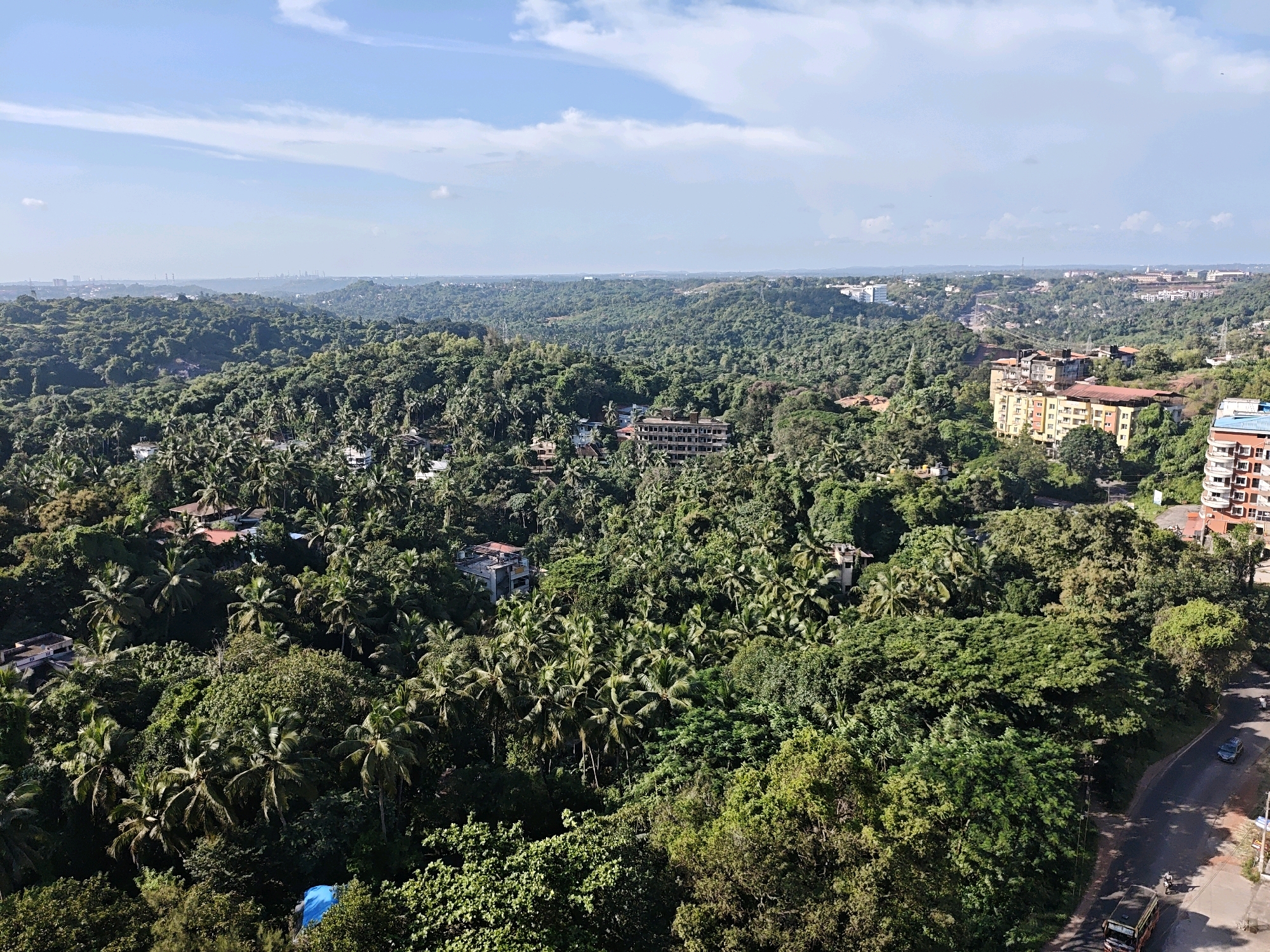 This is an aerial, high-angle, panoramic shot of Kudupu, India, on a clear, sunny day with a few scattered clouds. The foreground is dominated by a dense, lush green canopy of trees, predominantly palm trees, interspersed with other foliage. Several buildings are visible nestled within the greenery, ranging from single-family homes with red-tiled roofs to larger apartment buildings and what appear to be commercial structures. A road curves through the lower right portion of the image, with a dark-colored bus and a lighter-colored car traveling on it. The road also has visible painted lines and a sidewalk. In the distance, more buildings and a hazy cityscape are discernible, with hills and more vegetation stretching towards the horizon. The overall impression is one of a city integrated with nature, showcasing a dense tropical environment.