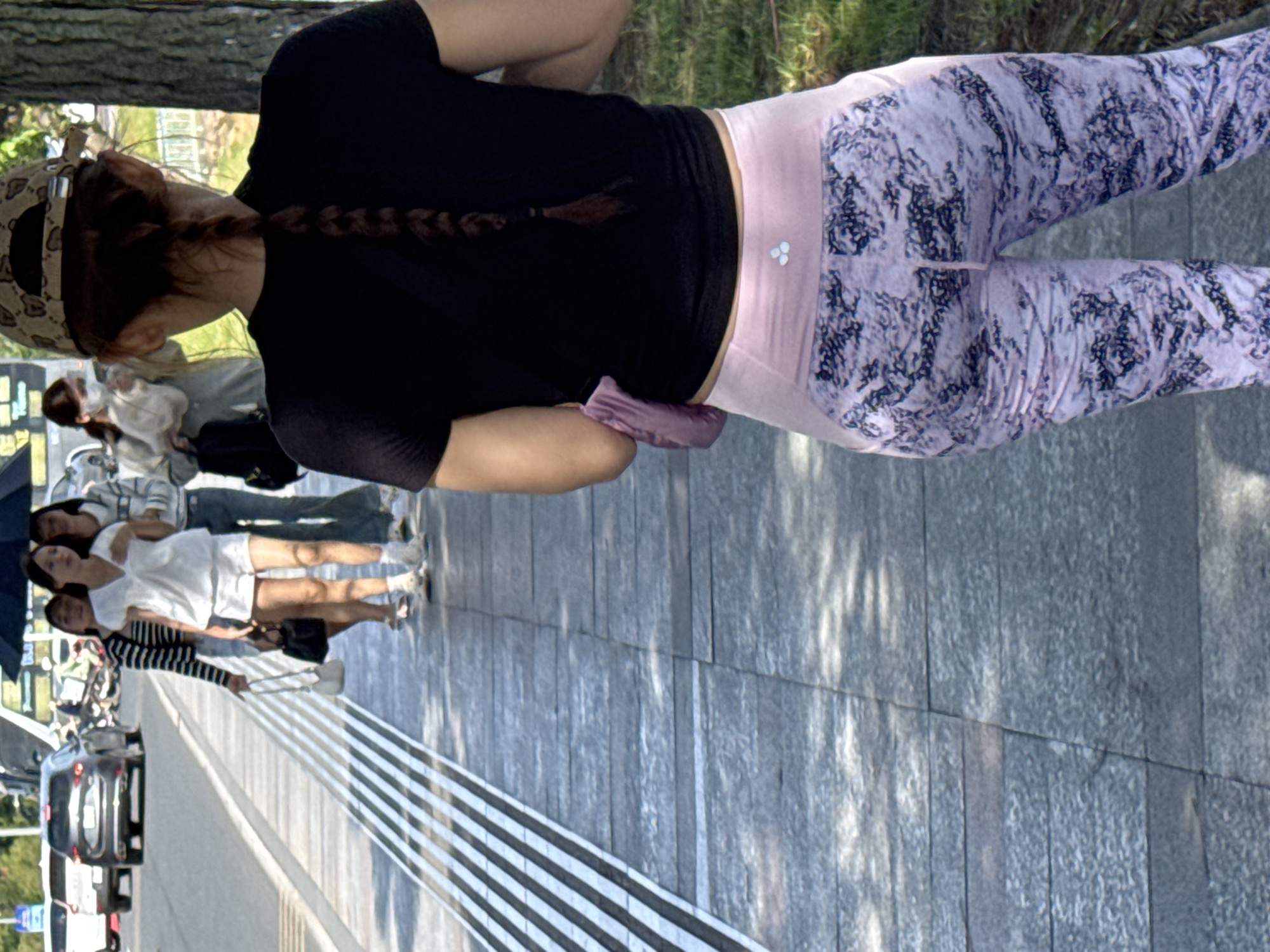 The image shows a street scene in Da Lat, Vietnam. In the foreground, a person with a braided brown ponytail and wearing a black t-shirt and patterned pink leggings is walking away from the camera. They have a light purple pouch or bag attached to their left hip. The background shows several other people walking on a sidewalk next to a street. One person is wearing a striped long-sleeved shirt and a white skirt, carrying a white handbag. Another person is in a white t-shirt and white shorts, also carrying a black handbag. Further back, a person in a light-colored sweater and jeans is visible, and another person in a light grey jacket is wearing a face mask. Several cars are parked and driving on the street, including a silver SUV. The environment appears to be a paved sidewalk with a row of raised tactile paving strips along the edge of the street. Trees are visible on the right side of the image. The lighting suggests it is daytime with dappled sunlight casting shadows on the sidewalk.