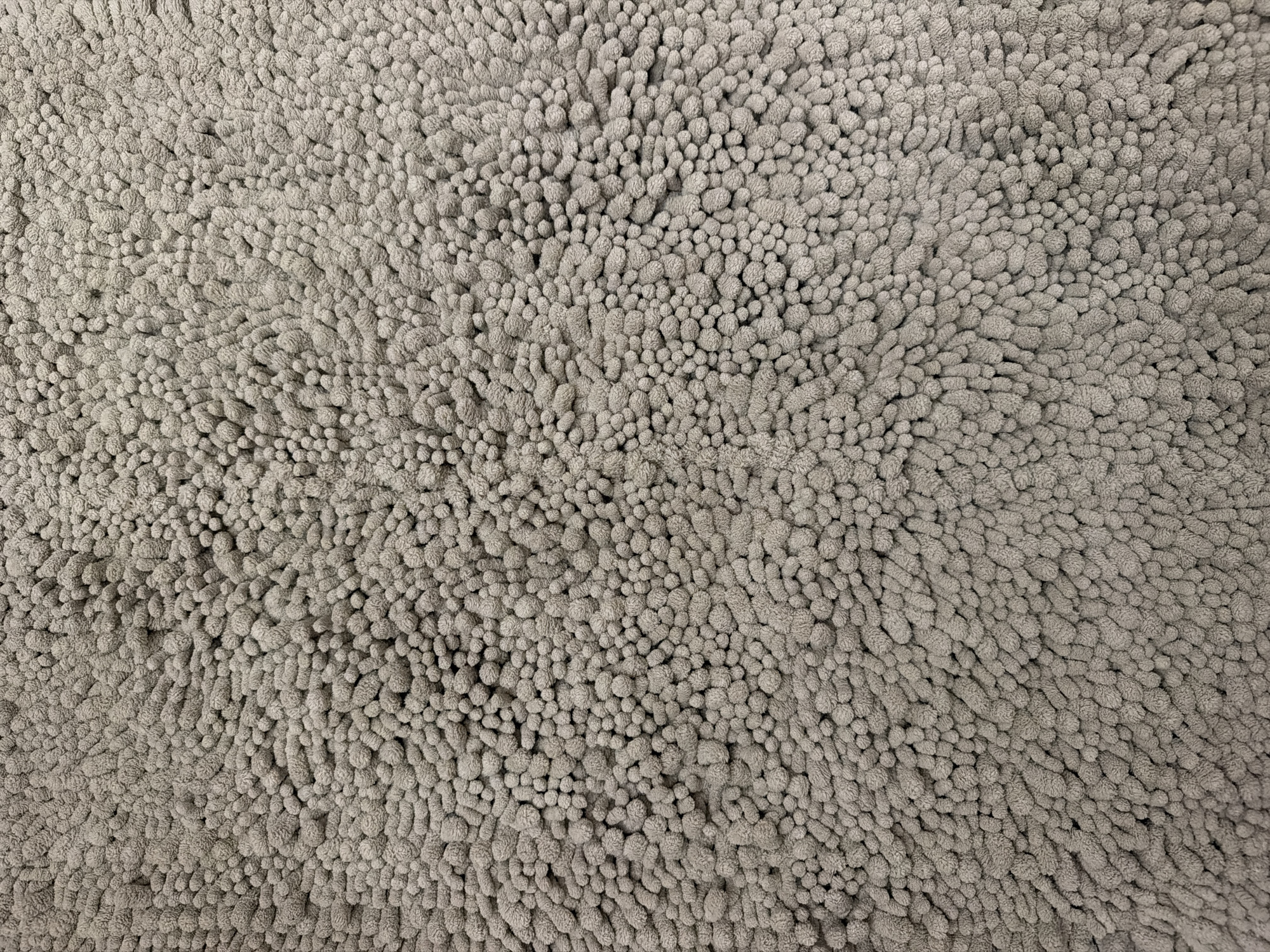 The image is a close-up, overhead view of a shaggy, grey carpet. The carpet is made of numerous small, tightly packed loops or shag fibers, giving it a plush and textured appearance. The fibers are all the same color, a neutral grey, and the lighting creates subtle shadows that highlight the depth and unevenness of the texture. There are no people, objects, or discernible features of a specific location visible. The image is a purely abstract depiction of carpet texture. Due to the abstract nature of the image, it is impossible to determine its location in the city of Khmelnytskyi, Ukraine.