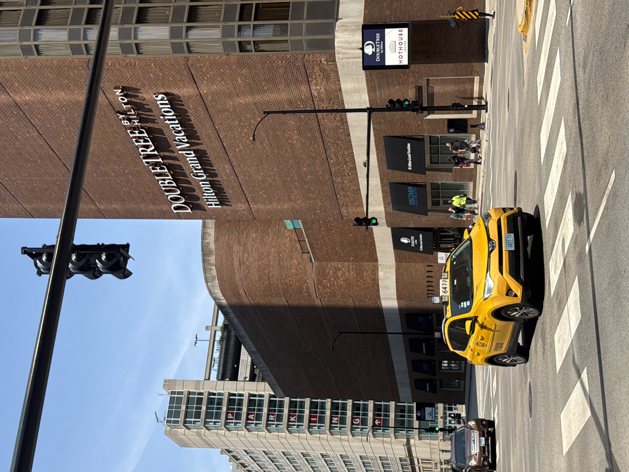 This image captures a street-level view of an intersection in Chicago, Illinois, USA, on a bright, sunny day. A yellow taxi, with the number "6477" visible on its roof, is in the foreground, waiting at a crosswalk. To the right of the taxi, a brown SUV is also proceeding through the intersection.
Dominating the background is a large, brick building with the prominent signage "DOUBLETREE BY HILTON" and "Hilton Grand Vacations." Adjacent to this is a taller, modern building with a visible "PARKING" sign spelled out vertically on its facade. Traffic lights hang over the intersection, with the green light illuminated for vehicles proceeding forward.
Several pedestrians are visible walking on the sidewalk in front of the Doubletree building. One person in a bright yellow safety vest stands out. A street lamp with a black pole is positioned near the sidewalk. The environment suggests an urban setting with clear blue skies overhead.