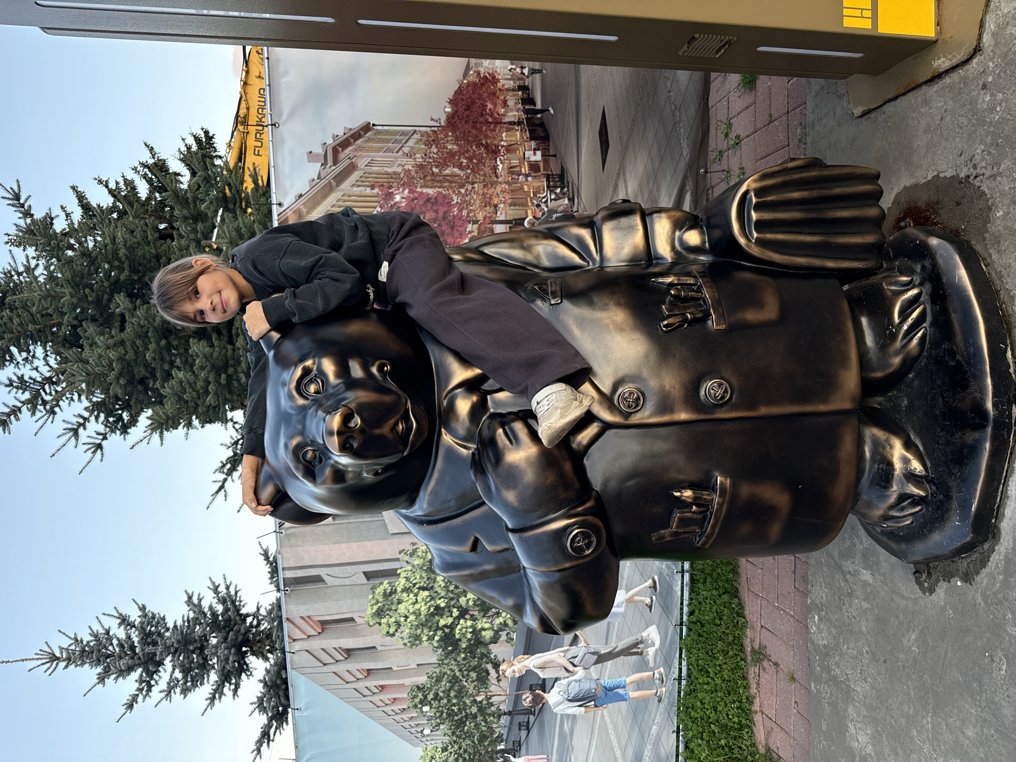 A young girl, appearing to be of East Asian descent, sits atop a large bronze statue of a bear dressed in a suit. The bear statue is positioned outdoors, in front of a large banner depicting a street scene in a city. The banner shows buildings, trees with red foliage, and people walking. The girl is wearing a dark grey sweatshirt and dark grey pants. She is looking towards the camera with a slight smile. Her right arm is resting on the bear's head, and her left leg is bent, with her foot resting on the bear's shoulder. The bear statue has its hands clasped in front of its chest. In the bear's left breast pocket, there are several art supplies, including pens and brushes. The bear is also holding a briefcase in its right hand. The ground in front of the statue is paved with bricks. There are trees and a blue sky in the background. The overall impression is of a playful interaction between a child and a public art installation. There is no information provided to suggest the image was taken in Rusanovo, Russia.