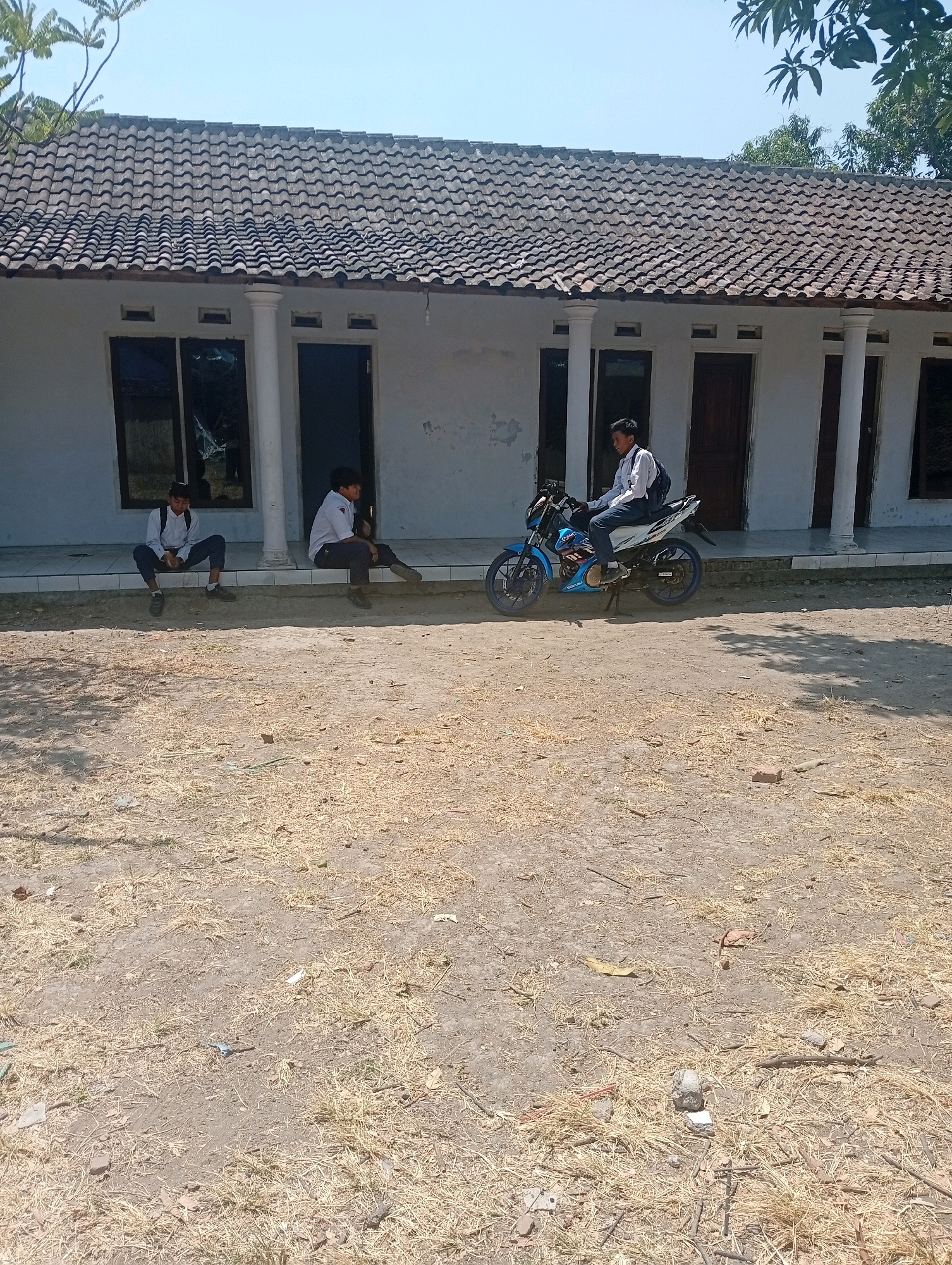In front of a white building with a tiled roof, two young males in school uniforms are seated on a low concrete ledge. One is positioned on the left, with his legs crossed and his hands resting on his knees. The other is to his right, facing away from the camera, with his legs extended. To the right of these two individuals, a third young male, also in school uniform with a backpack, sits on a blue and white motorcycle. The motorcycle is parked on a dry, dusty ground with scattered straw and small debris. The building has multiple doors and windows, with white columns supporting the roof overhang. Trees with green leaves are visible in the background under a clear blue sky. The scene appears to be outdoors, likely in a school or community area in Gempeng, Indonesia, during daylight hours.