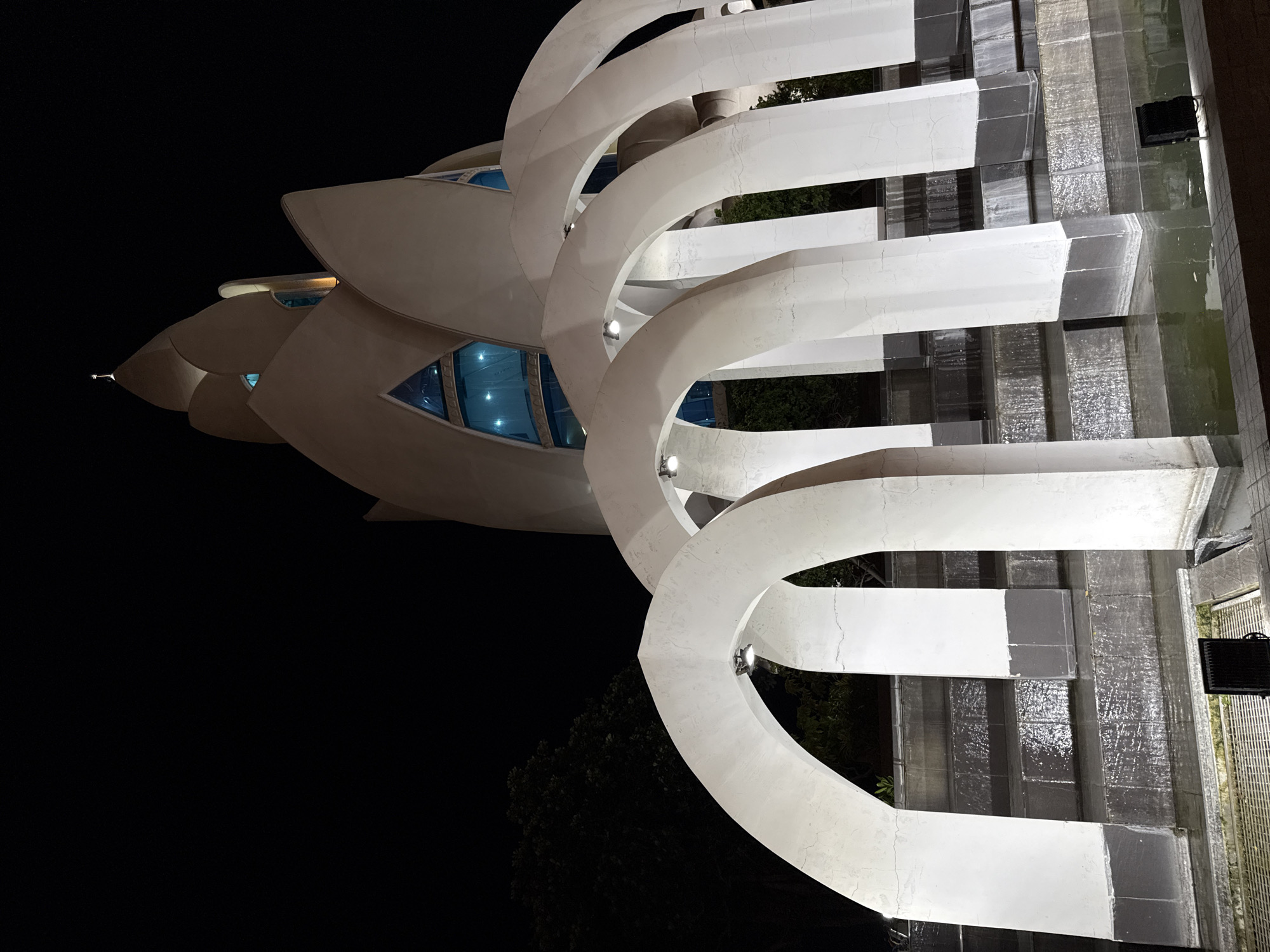 The image depicts the Long Son Pagoda in Nha Trang, Vietnam, at night. The foreground features a series of white, arched structures that appear to be part of a water feature or fountain, with water cascading down tiered levels. The arches are illuminated by spotlights. Behind the arches, the main structure of the pagoda is visible, characterized by its unique lotus-shaped roof and white, curved facade. Blue-tinted windows are set into the pagoda's design. A cross is visible at the apex of the pagoda's tallest point. The background is dominated by a dark, clear night sky. The scene is illuminated by artificial lighting, highlighting the architectural features of the pagoda and the water feature. There are no people visible in the image.