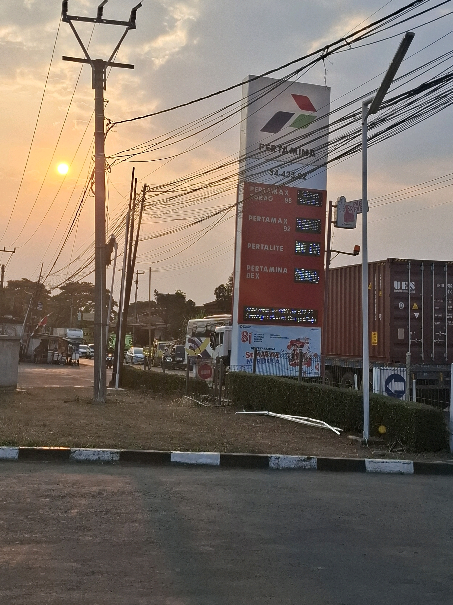 The image depicts a Pertamina gas station sign in Karangtengah, Indonesia, with a setting sun visible in the upper left quadrant. The sign displays prices for various fuel types including Pertamax Turbo, Pertamax, Pertalite, and Pertamina Dex, with prices listed next to each. The sign is tall and rectangular, illuminated with digital displays for the prices. Overhead, a complex network of electrical wires crisscrosses the sky. To the right of the sign, a shipping container is visible. In the foreground, the road is paved, and a curb with black and white markings runs along the edge of the road. The overall scene suggests a typical urban or suburban environment with active traffic, indicated by several vehicles in the background, including a bus and a truck. The lighting suggests it is late afternoon or early evening.