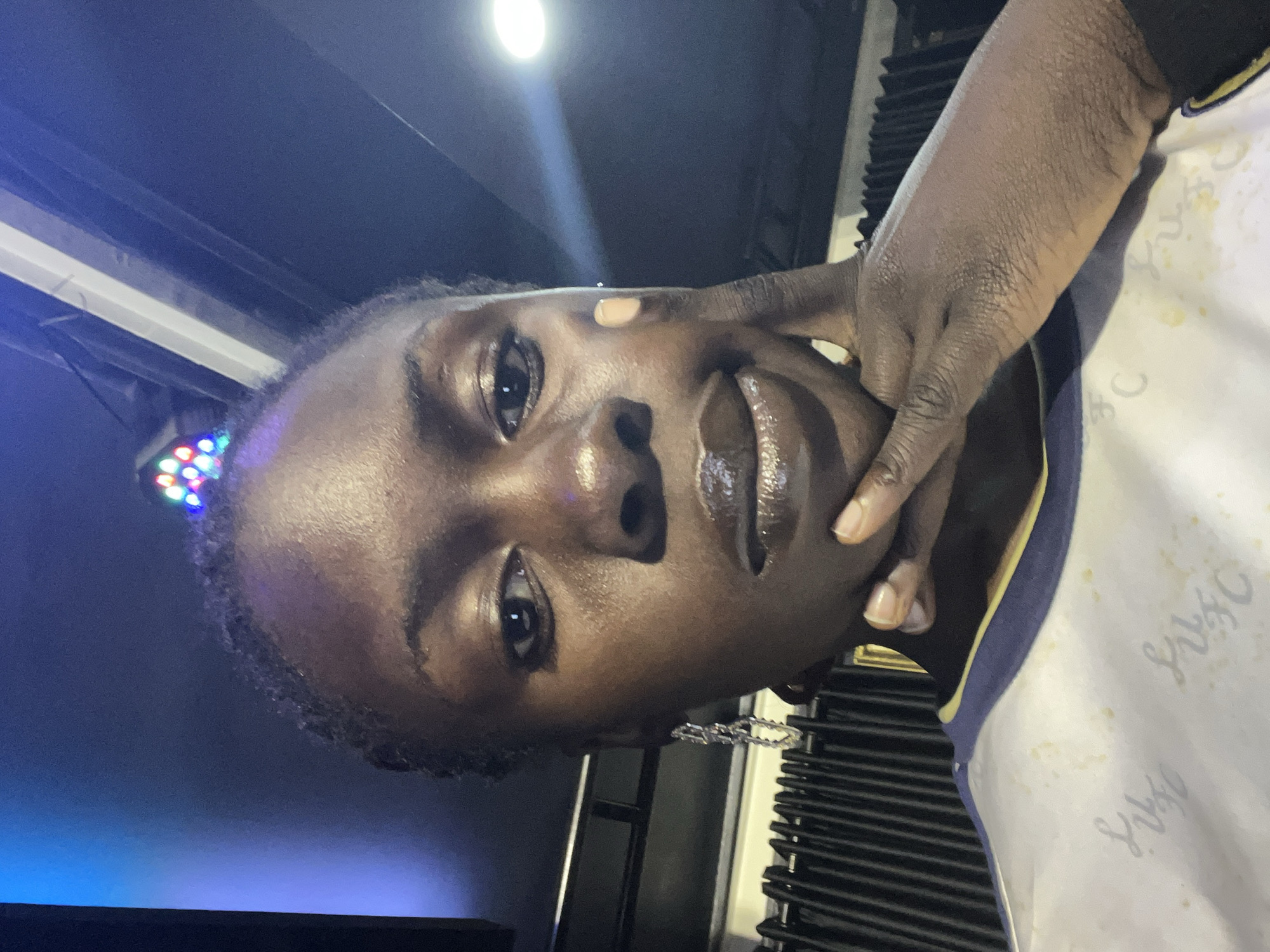 A close-up, indoor selfie depicts a young Black woman from the chest up, with her face occupying the majority of the frame. She has dark skin and her hair is styled in braids, adorned with small, colorful lights that are illuminated. She is looking directly at the camera with a neutral expression. Her right hand is resting on her chin and cheek, and her left hand is also visible near her face. She is wearing a white t-shirt with a blue and yellow collar, and a silver, dangling earring on her left ear. The background is dark and appears to be an indoor venue with a ribbed black curtain behind her and blue and white lighting above. There is a bright, circular light source visible in the upper right portion of the image. The image does not contain any elements that would allow for specific identification of the location within Jos, Nigeria.
