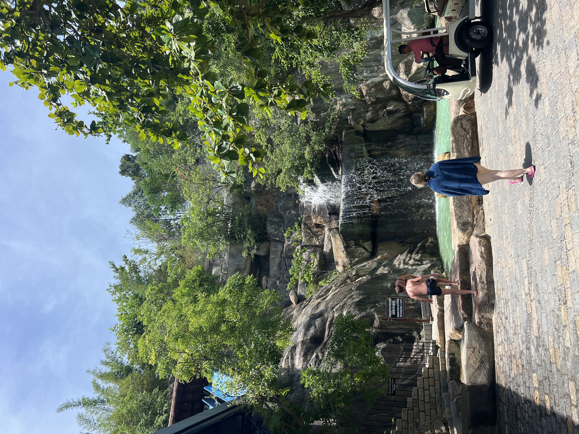 This is a medium shot of a man in a pink shirt driving a golf cart in Tay Nha Trang Ward, Vietnam. To his left, a woman in a dark blue cover-up and pink flip-flops walks away from the camera towards a waterfall. Two boys in swim trunks are walking on large stone steps leading to the waterfall and a turquoise pool. The waterfall is cascading down a rocky cliff face surrounded by lush green trees. A sign with Vietnamese text is visible on the rock face.