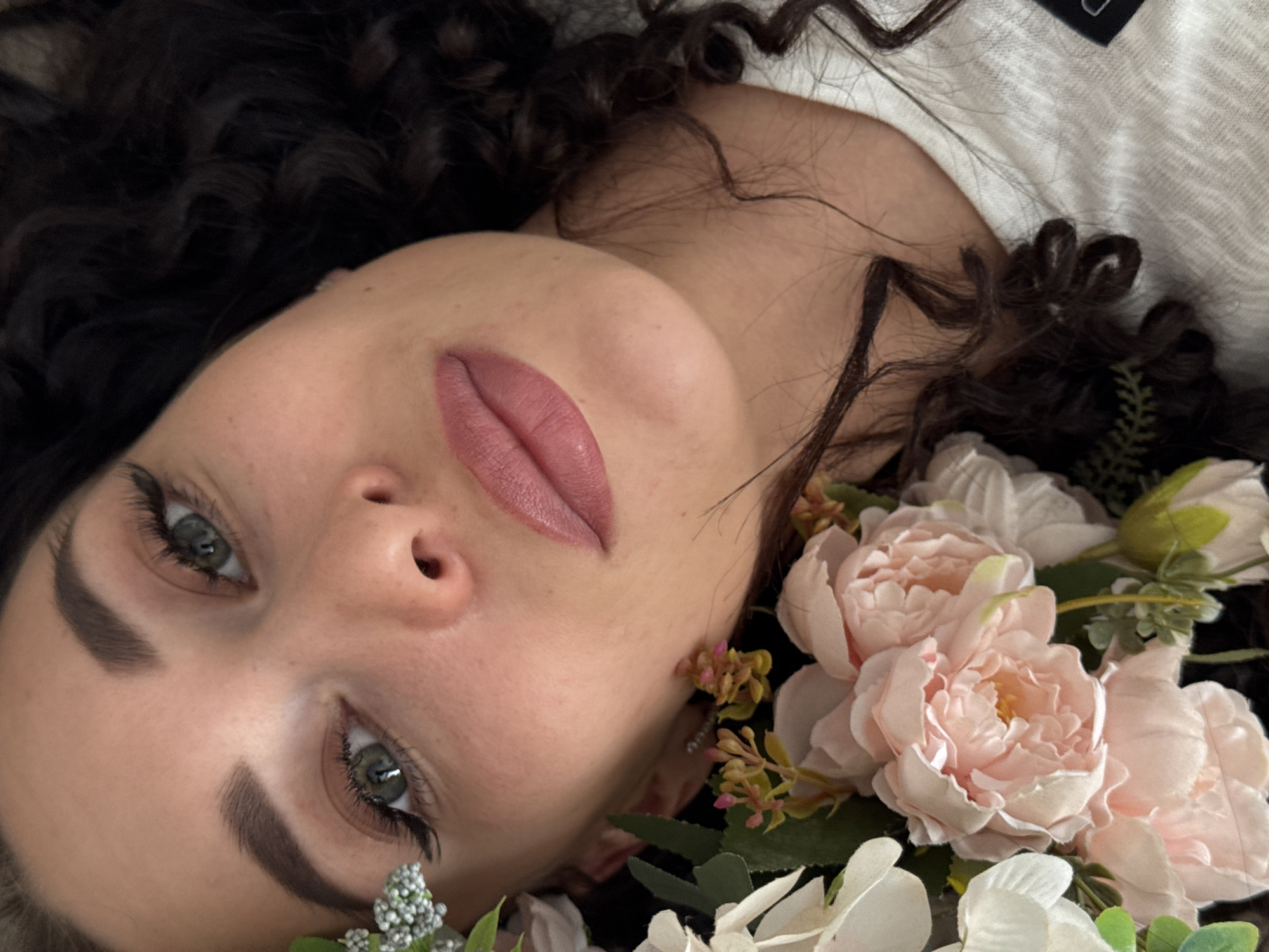 A close-up, tilted shot depicts a woman with dark, curly hair lying down among light pink and white flowers with green foliage. Her face is angled towards the camera, showcasing her fair skin, dark eyebrows, green eyes, and pink lips. She is wearing what appears to be a light-colored garment. The background is indistinct, but suggests an indoor setting. There is no indication of the city of Chaikovsky, Russia.