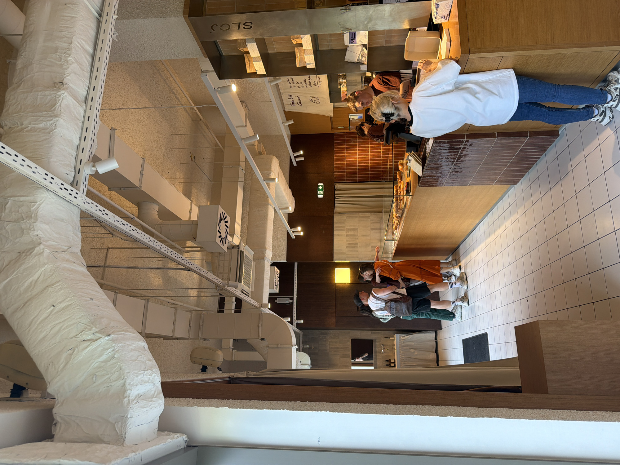The image depicts the interior of a bakery or cafe in Belgrade, Serbia, named "Sloj." A female customer in a white shirt and jeans is paying at the counter. Another customer, wearing an orange top, is also present with an acquaintance. The interior features a display case of pastries, a wooden counter, and tiled flooring. The ceiling has exposed ductwork and industrial lighting. Signage with "Sloj" is visible above the counter.
