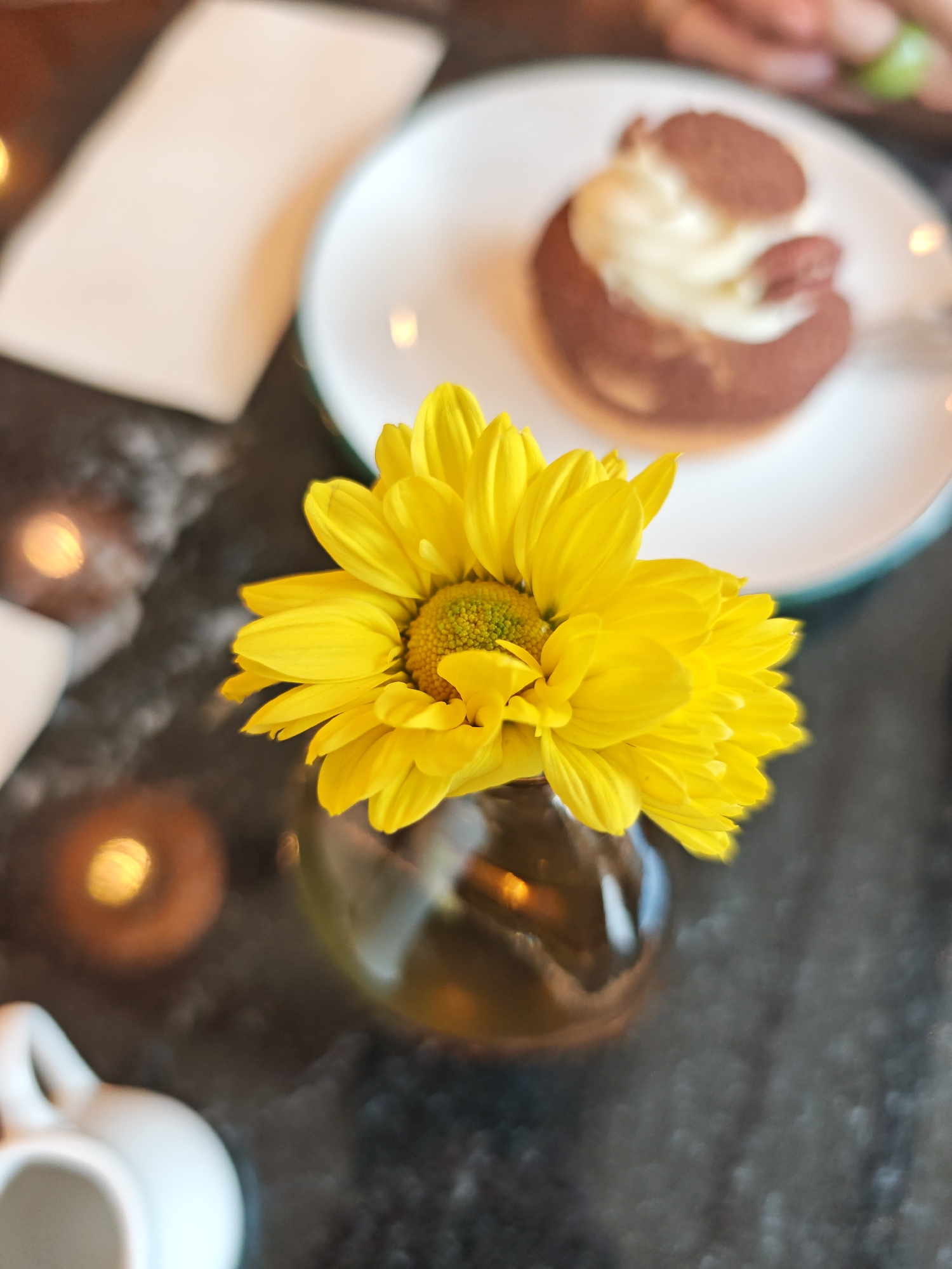 A bright yellow daisy is arranged in a small amber glass vase, positioned centrally in the frame. The flower's petals are in sharp focus, with its vibrant yellow color contrasting against the blurred background. To the right of the flower, a white plate holds a dessert topped with whipped cream and dusted with cocoa powder. In the upper left, a white napkin is visible. The surface beneath the objects appears to be a dark, textured table, possibly granite, with bokeh effects suggesting ambient lighting or out-of-focus elements. In the bottom left corner, a portion of a small white creamer is visible. The overall scene evokes a cozy café atmosphere. No specific location in Sofia, Bulgaria, can be identified from the image.