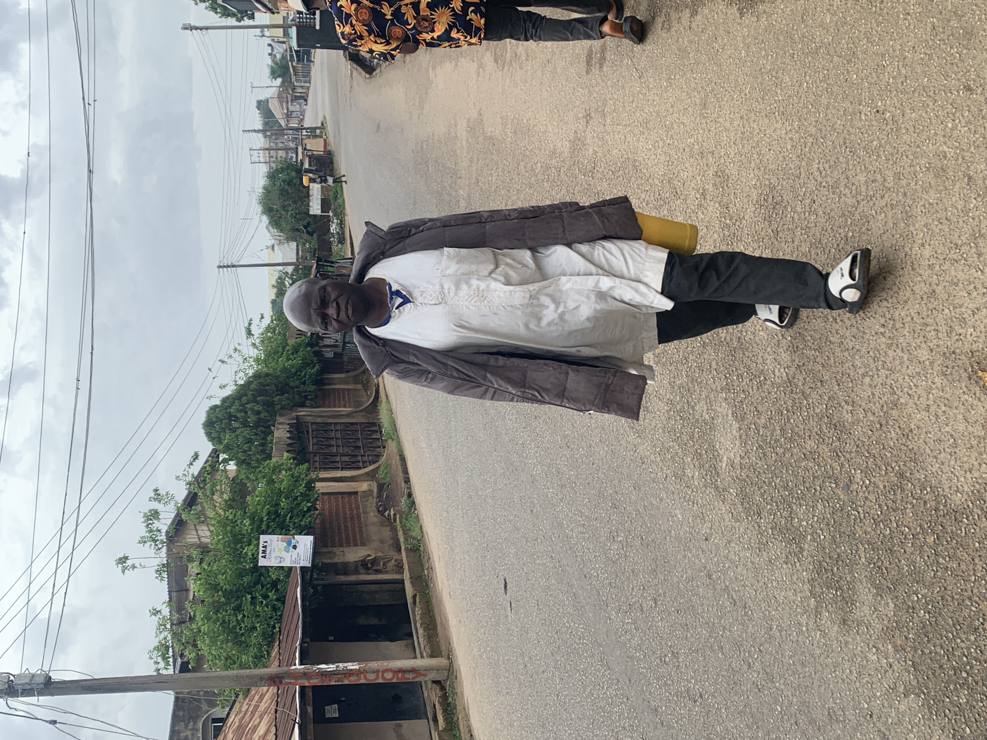A man stands in the middle of a paved road. He is wearing a dark-colored jacket, a white shirt, and black pants. He also has on white and black shoes. A yellow object is tucked under his arm. In the background, there are buildings on the left side of the road, partially obscured by vegetation. Overhead power lines run along the side of the road. In the distance, other buildings are visible. The sky is overcast. The image is taken at street level and has been rotated to the side. The buildings on the left of the image contain a sign for "AMA's". A possible location is Jos, Nigeria.