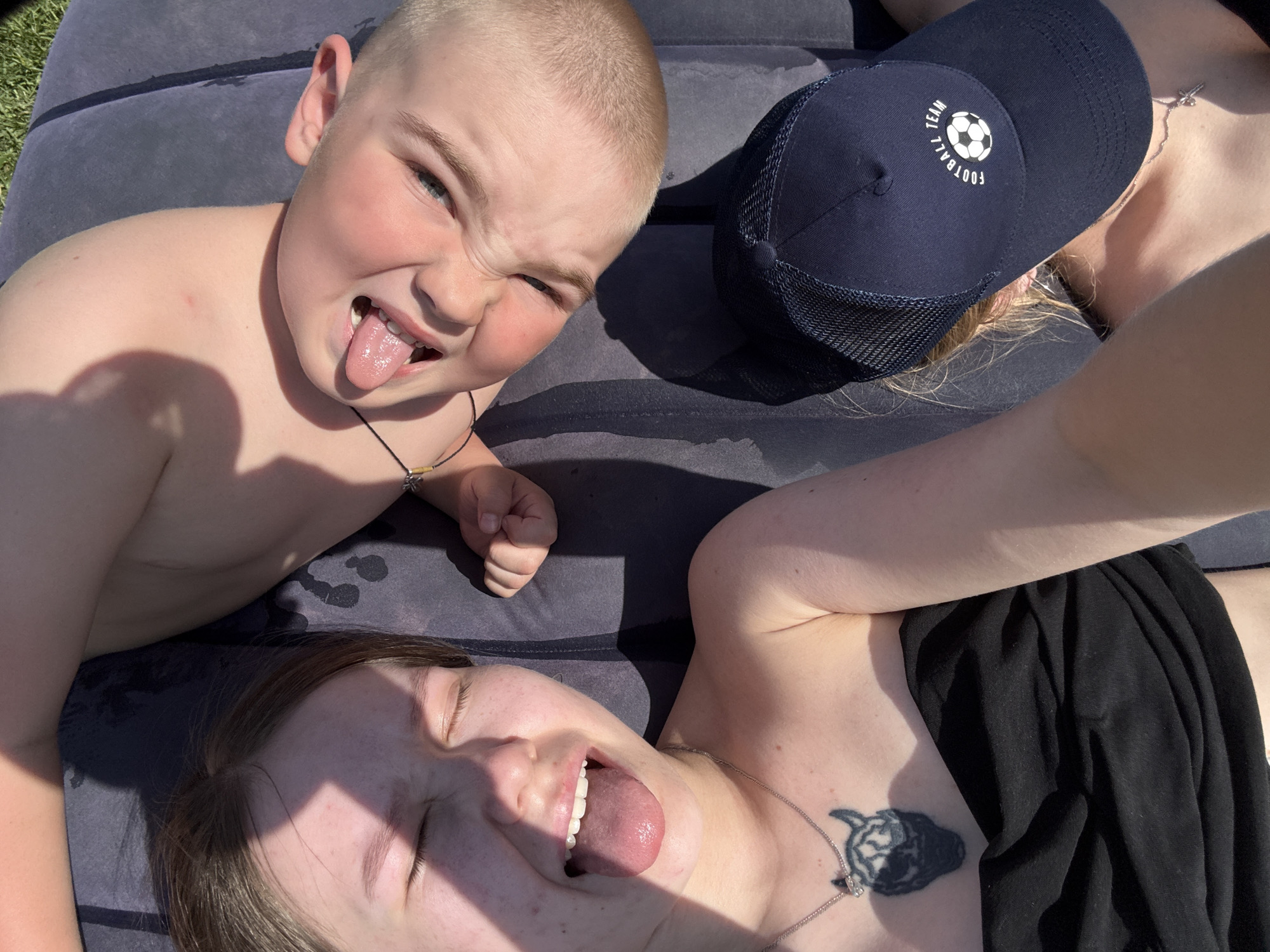A selfie shows three people lying on a dark blue inflatable raft outdoors in bright sunlight. A young boy with short, blond hair is in the upper left, sticking out his tongue and squinting. A woman in the lower left also sticks out her tongue, with her eyes closed. The third person wears a dark blue baseball cap with a white logo and a mesh back. The woman has a tattoo on her upper chest and wears a silver necklace. She is also wearing a black top. The raft appears to be slightly wet. The location is Zhigalki, Russia.